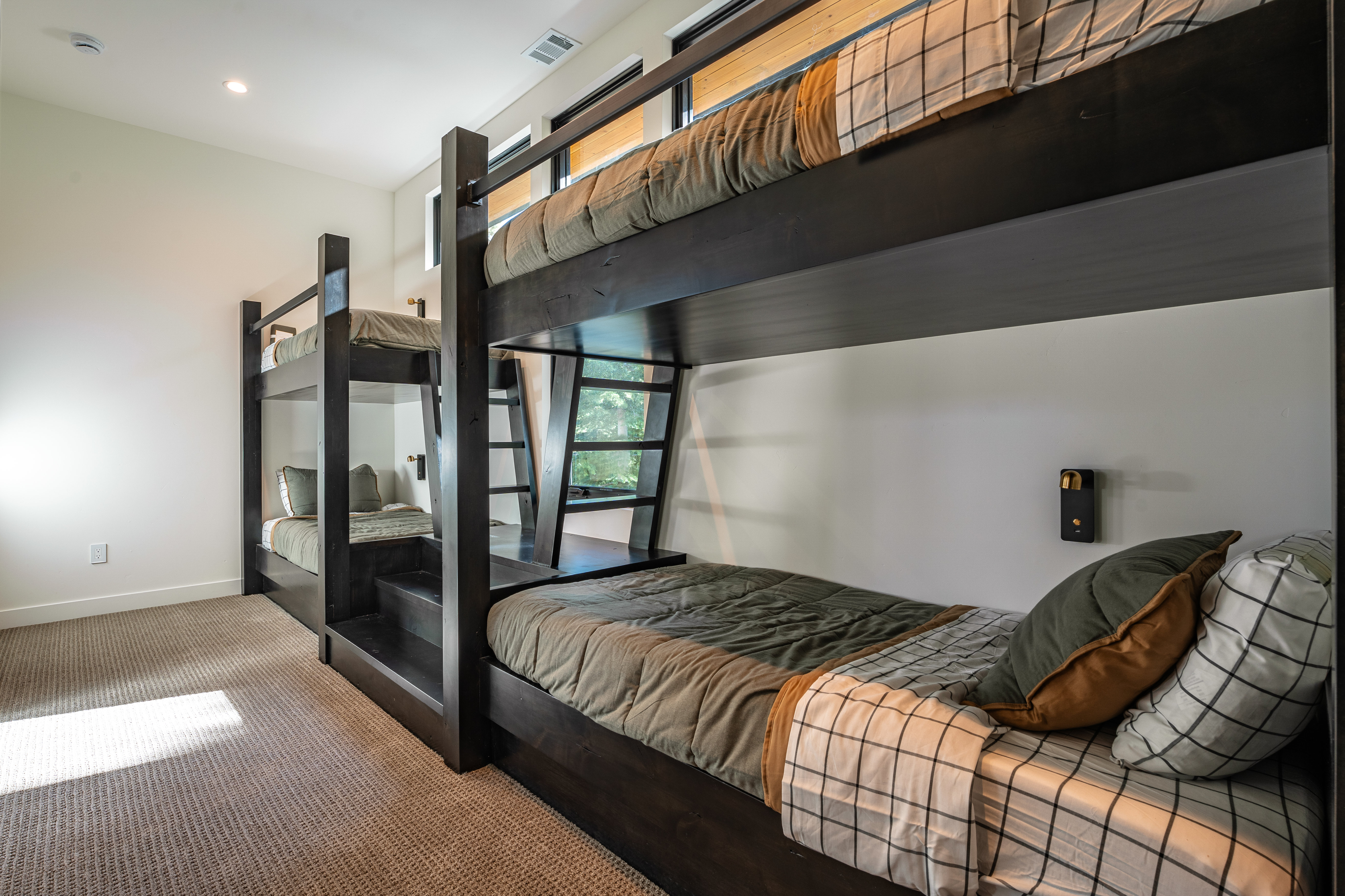This is a bedroom featuring two sets of dark wood bunk beds with neutral-toned bedding and plaid accents. The room has light-colored walls, carpeted flooring, and small windows near the ceiling, creating a cozy and functional space. A unique wall-mounted light fixture adds a touch of modern design.
