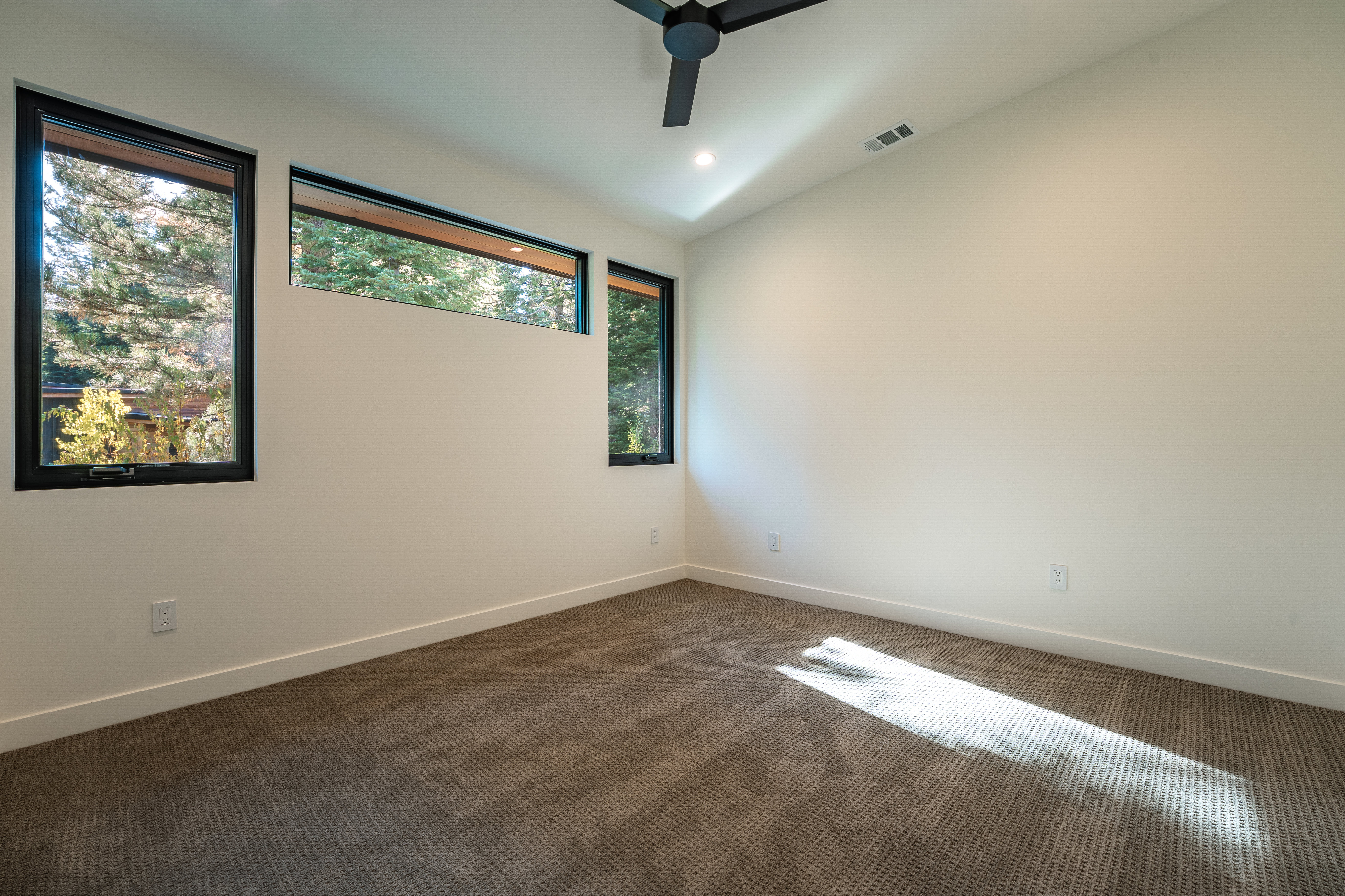 This is a well-lit, modern bedroom featuring neutral-toned walls and carpet. Three windows provide natural light and views of the surrounding trees. A ceiling fan is present, and the room appears spacious and uncluttered, offering a blank canvas for personalization.
