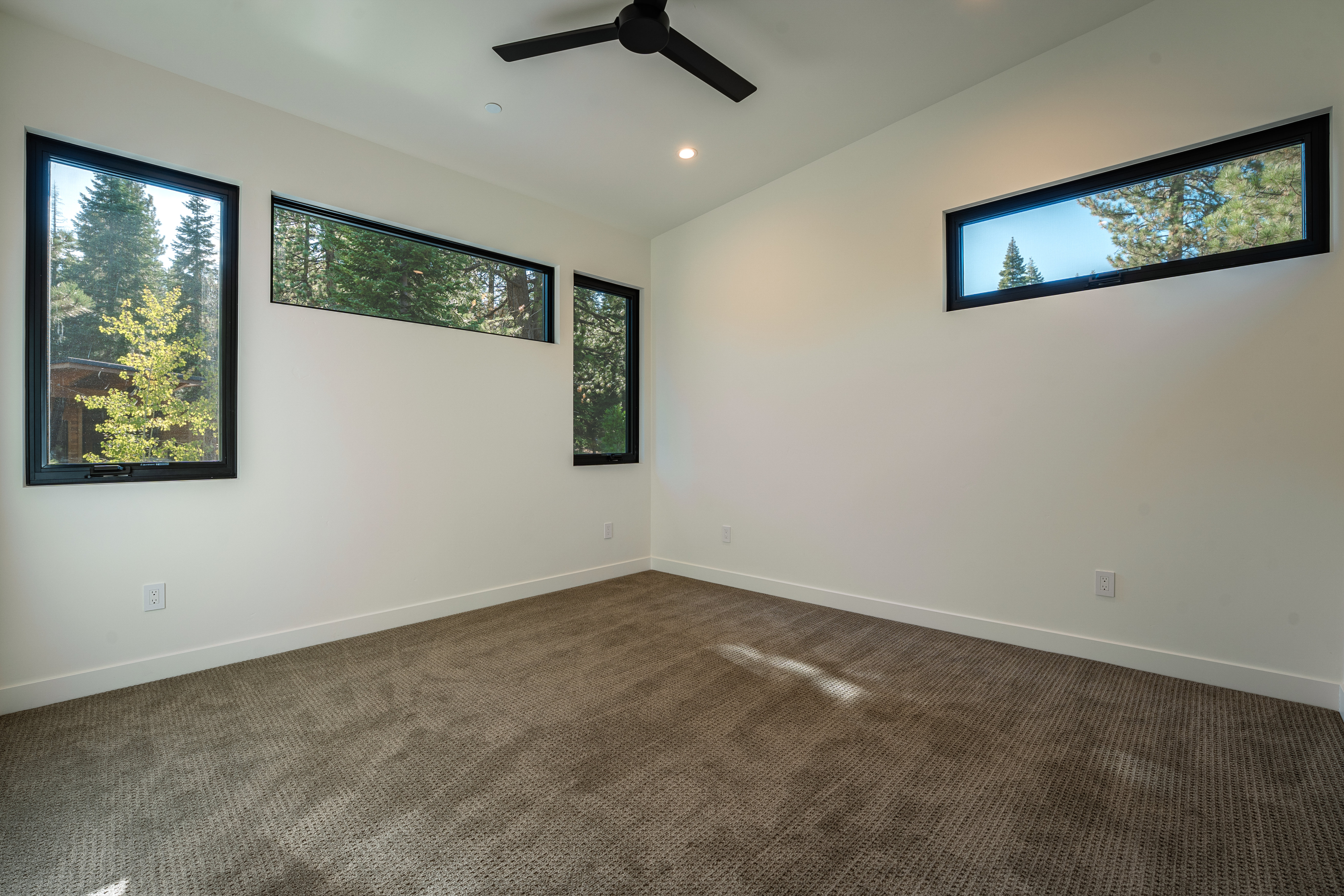 This is a bright, modern bedroom featuring neutral-toned carpet and white walls. Three windows with black frames offer views of the surrounding trees, enhancing the room's connection to nature. A black ceiling fan adds a contemporary touch, and the overall impression is clean and minimalist.
