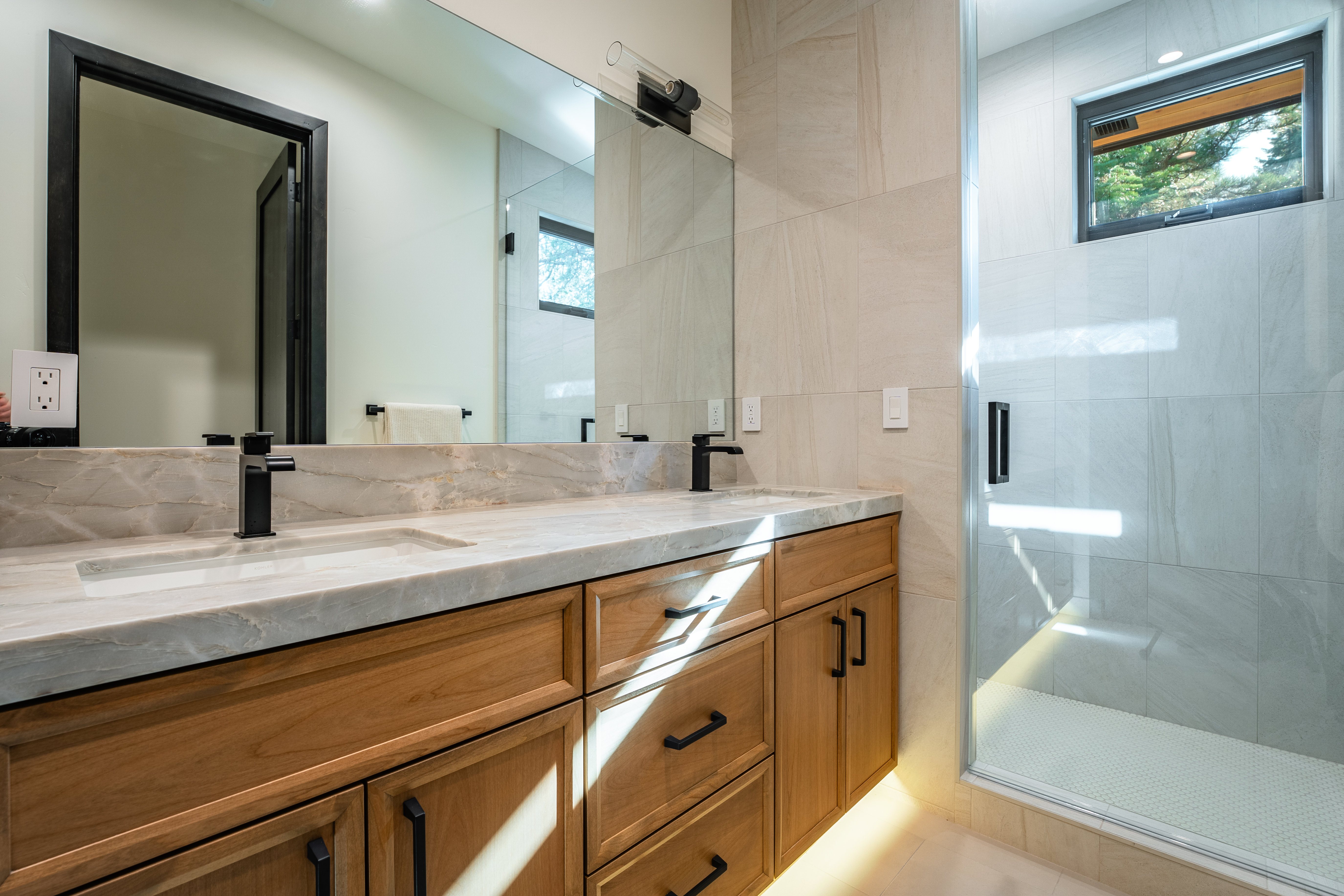 This is a well-lit primary bathroom featuring a double vanity with a marble countertop and wooden cabinets with black hardware. A large mirror reflects the space, including a glimpse into another room, and a glass-enclosed shower is visible on the right. The bathroom has a modern and clean aesthetic with neutral-toned tiles and fixtures.