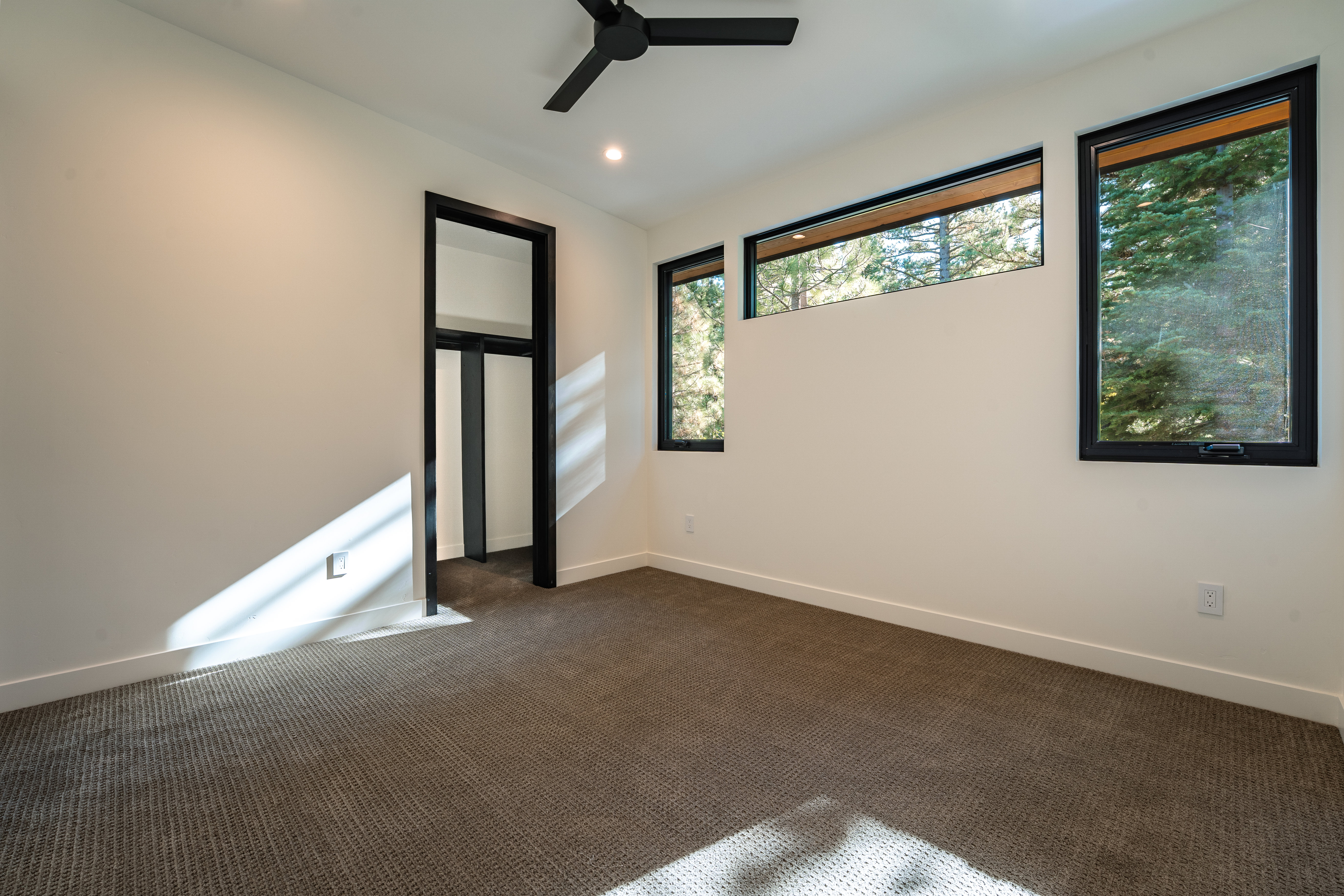 The image showcases a modern bedroom with neutral tones and clean lines. The room features a dark-framed closet, multiple windows offering natural light, and a ceiling fan. The carpet is a neutral color, and the walls are painted in a light shade, creating a bright and airy atmosphere.