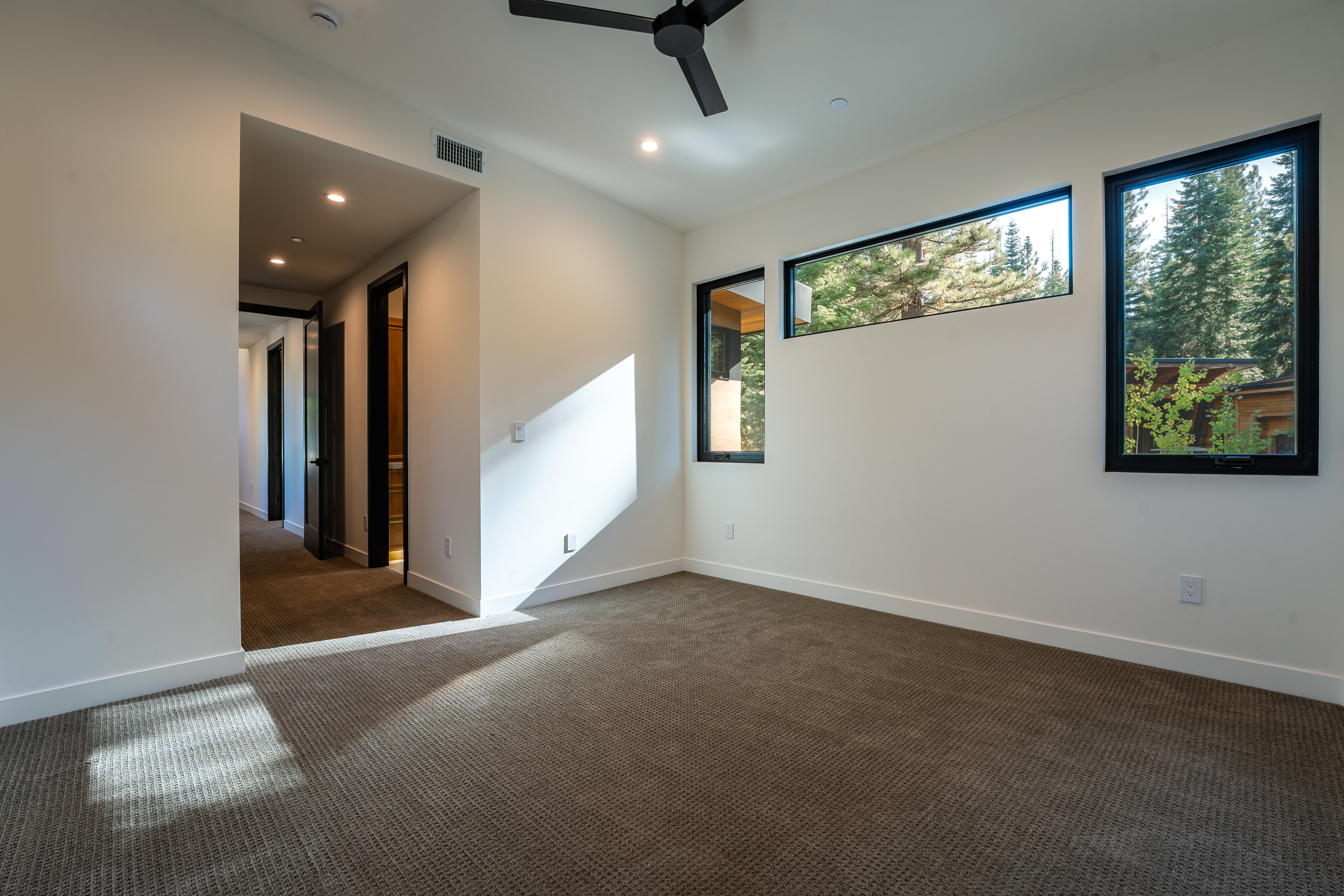 This is a spacious primary bedroom featuring neutral-toned carpet and white walls, creating a bright and airy atmosphere. The room has multiple windows offering natural light and views of the surrounding trees. A modern ceiling fan and recessed lighting provide additional illumination, while a hallway leads to other areas of the home.