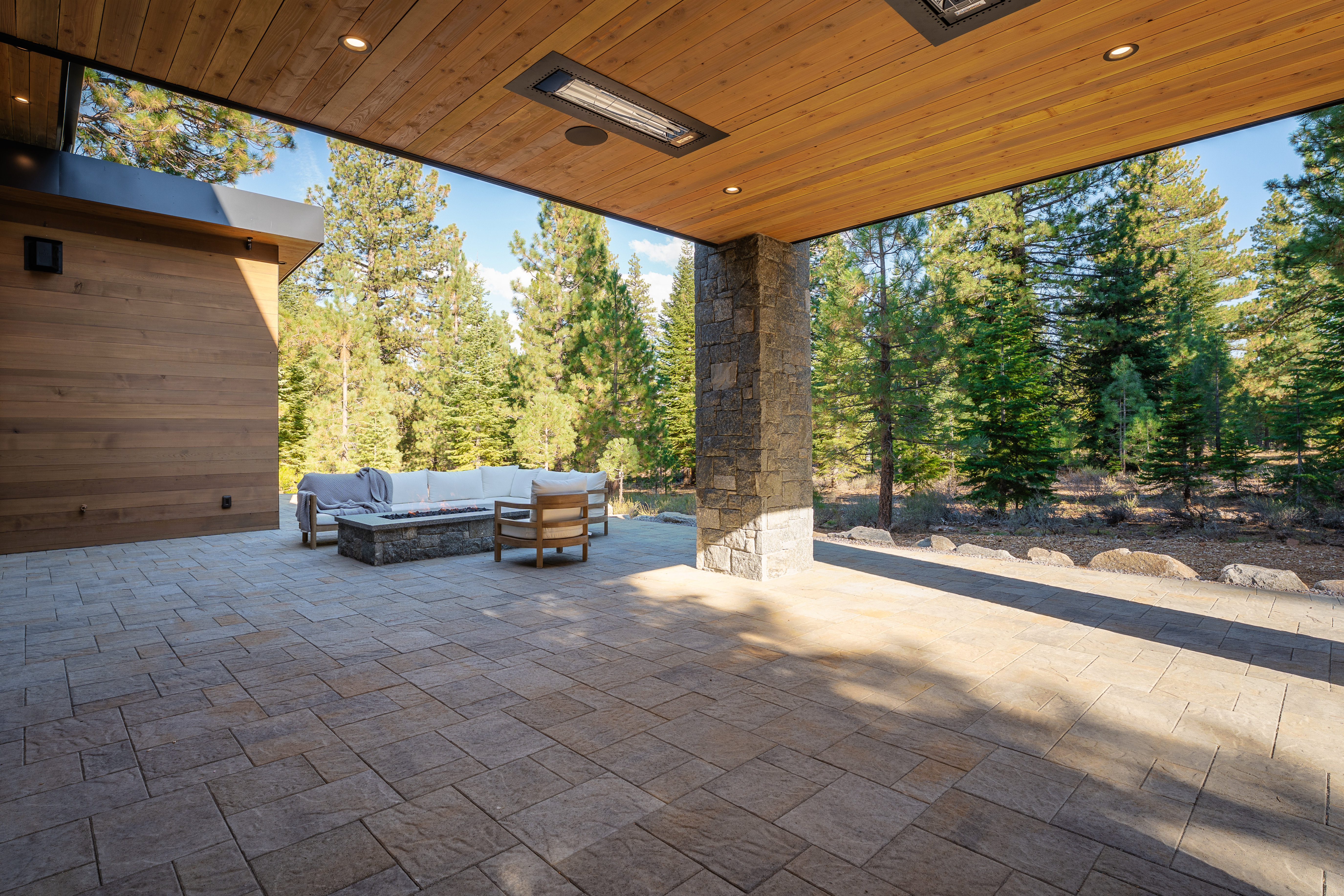 This image showcases a beautifully designed outdoor patio area. The patio features a stone-paved surface, a modern fire pit surrounded by comfortable seating, and a sturdy stone pillar supporting a wooden ceiling with recessed lighting. The space is framed by lush green trees, creating a serene and inviting atmosphere, perfect for relaxation and entertaining.
