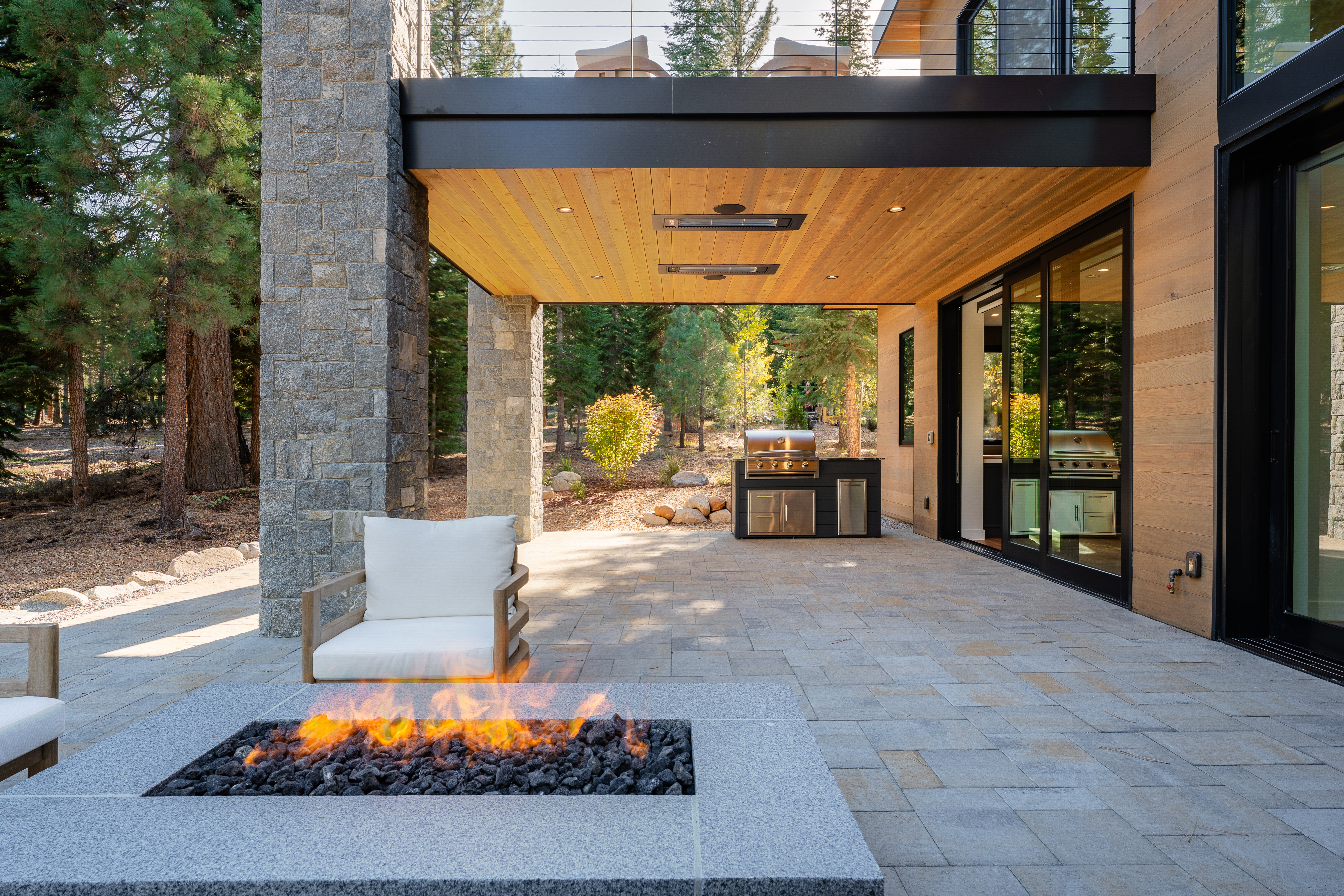 This image showcases a luxurious outdoor patio area, complete with a modern fire pit, comfortable seating, and a built-in outdoor kitchen with a stainless steel grill. The patio is paved with stone tiles and is partially covered by an overhang with wood paneling and recessed lighting. The space seamlessly blends indoor and outdoor living, offering a perfect setting for relaxation and entertainment.