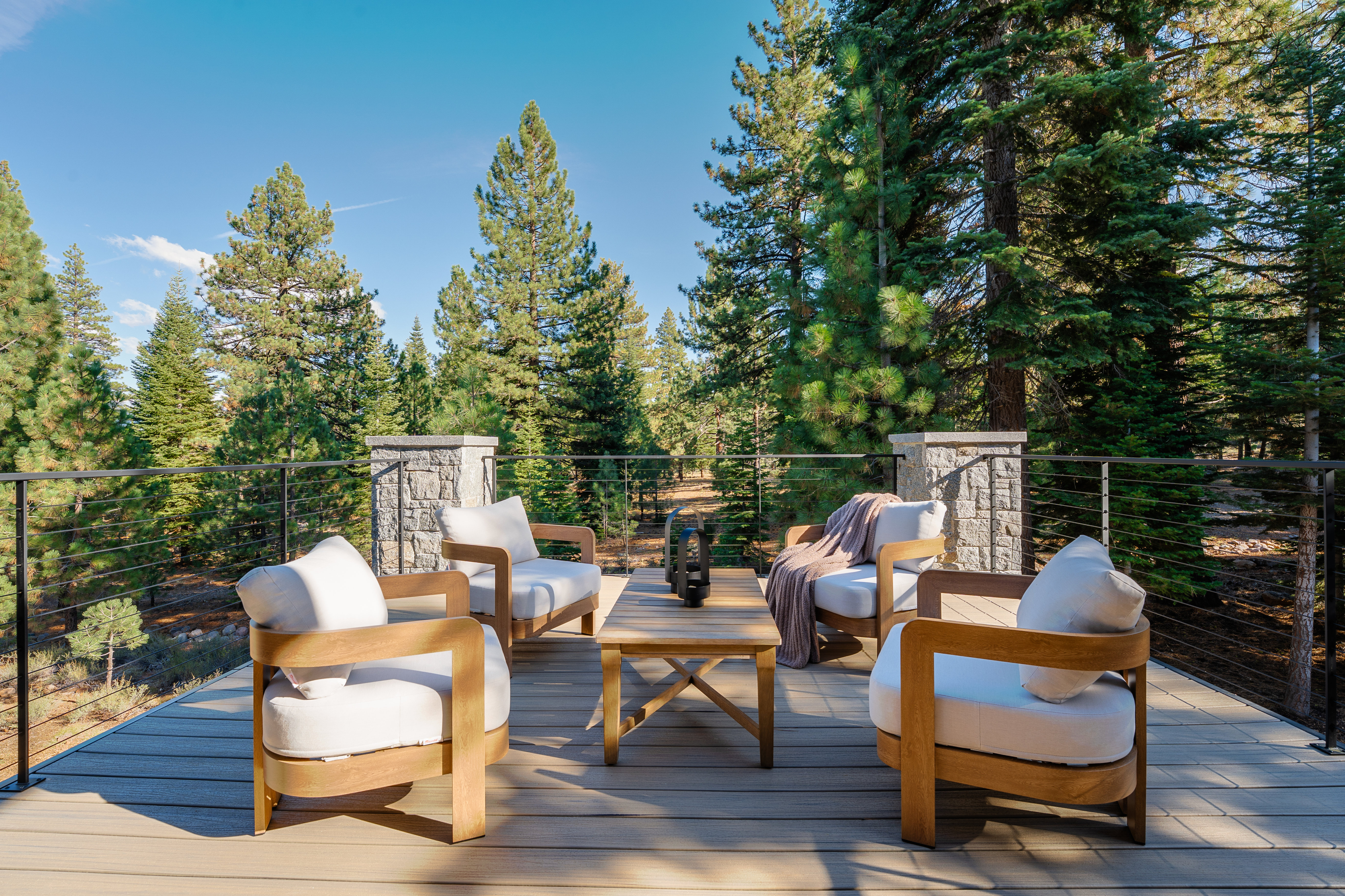 This image showcases a beautifully arranged outdoor patio or deck area, featuring four comfortable armchairs with white cushions surrounding a wooden coffee table. The deck is constructed with light-colored composite decking, and a modern railing system provides safety without obstructing the scenic view of the surrounding forest. Stone pillars add a touch of elegance and permanence to the structure.