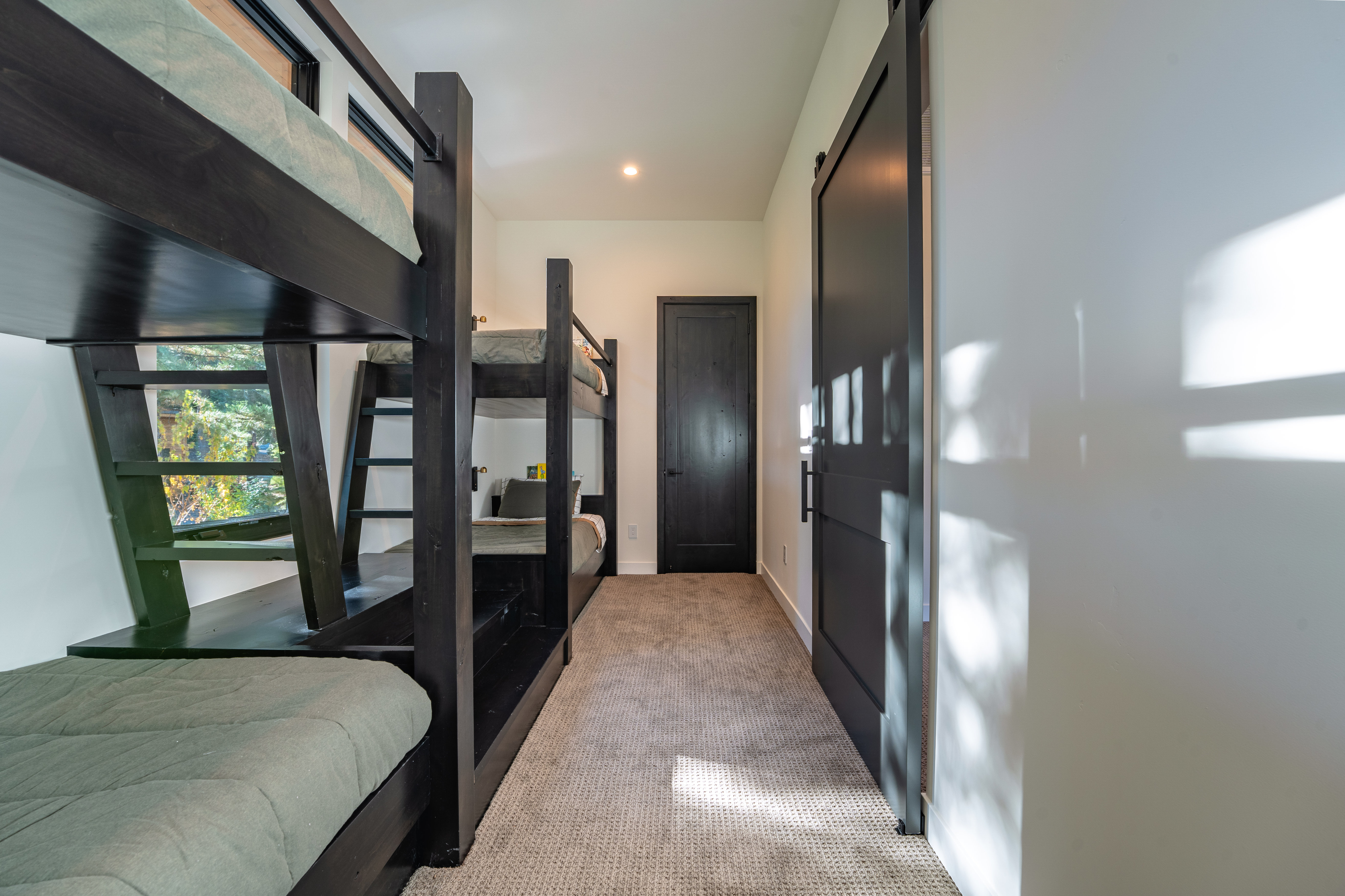 This interior shot showcases a bedroom featuring built-in bunk beds with a dark wood finish, creating a cozy and efficient use of space. The room is carpeted and includes a dark-colored door and a sliding barn door, adding a modern touch. Natural light filters into the room, highlighting the clean, neutral walls and the overall inviting atmosphere.