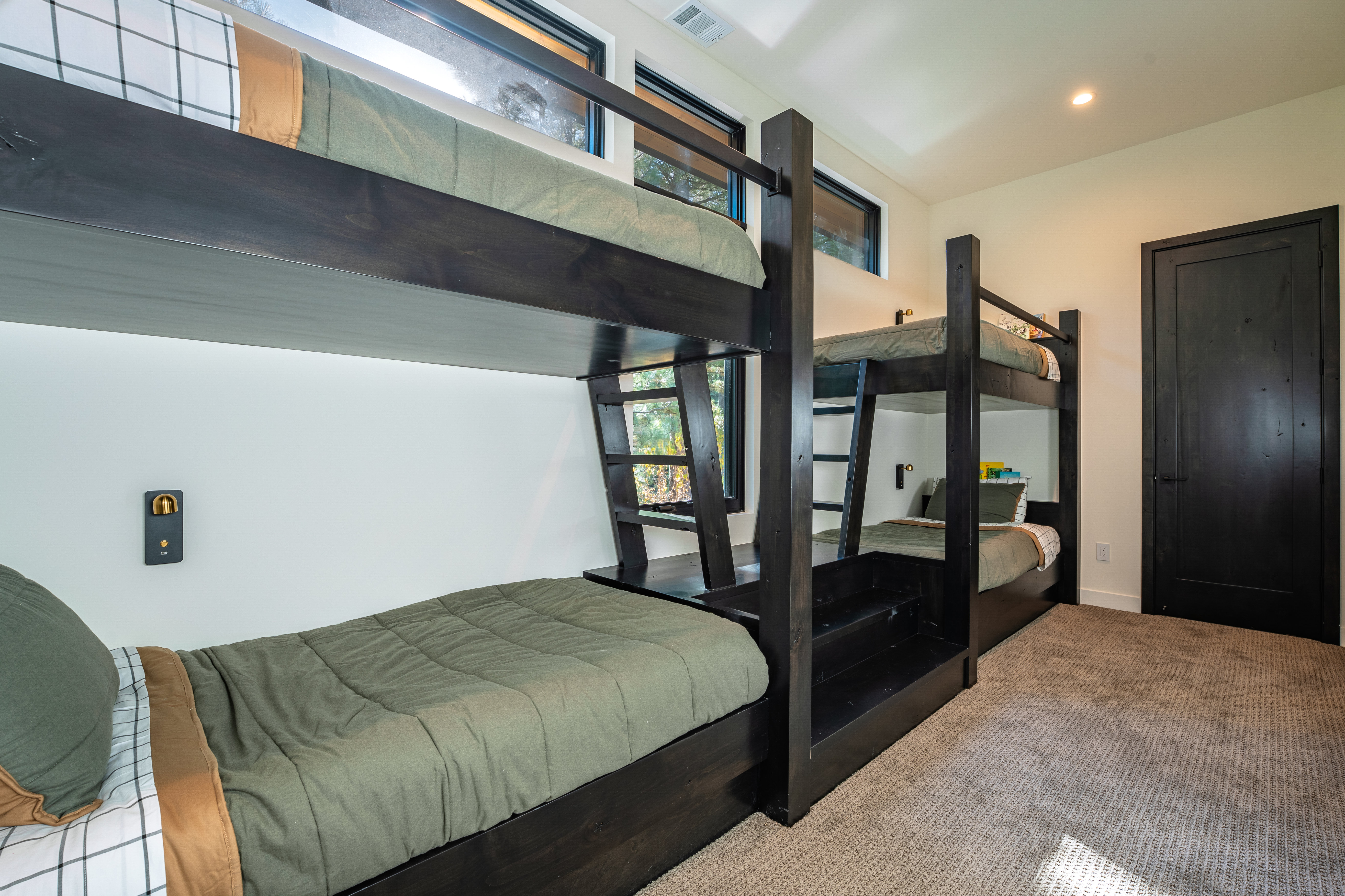 This is a bedroom featuring two sets of dark wood bunk beds, creating a cozy and functional space. The room has neutral-toned walls and carpet, with natural light coming in from the windows. The bedding is a muted green, contributing to a calm and inviting atmosphere.