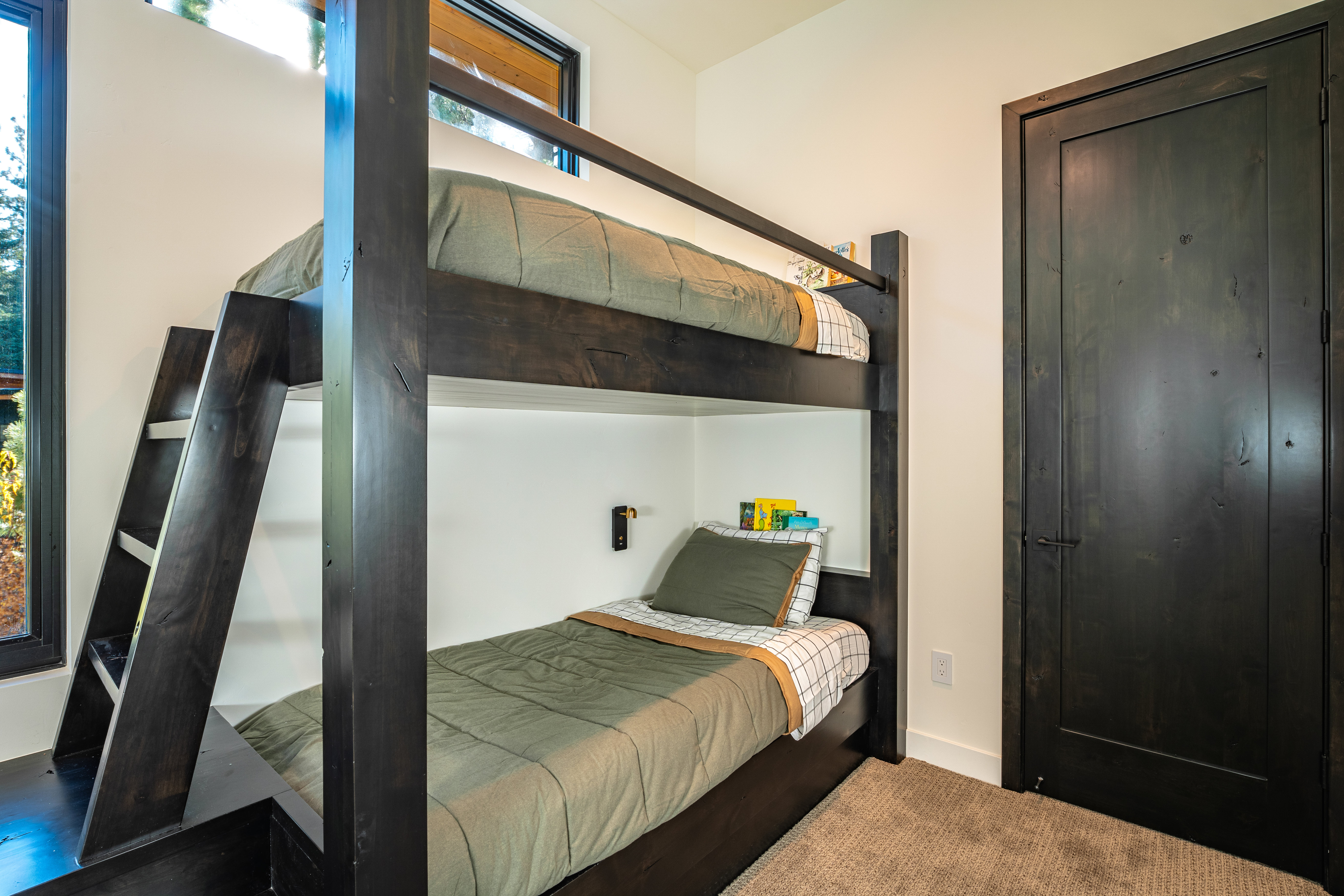 This is a cozy bedroom featuring a dark wood bunk bed with green bedding. The room has white walls and a dark wood door, creating a modern rustic feel. Natural light streams in from a nearby window, highlighting the clean and simple design.