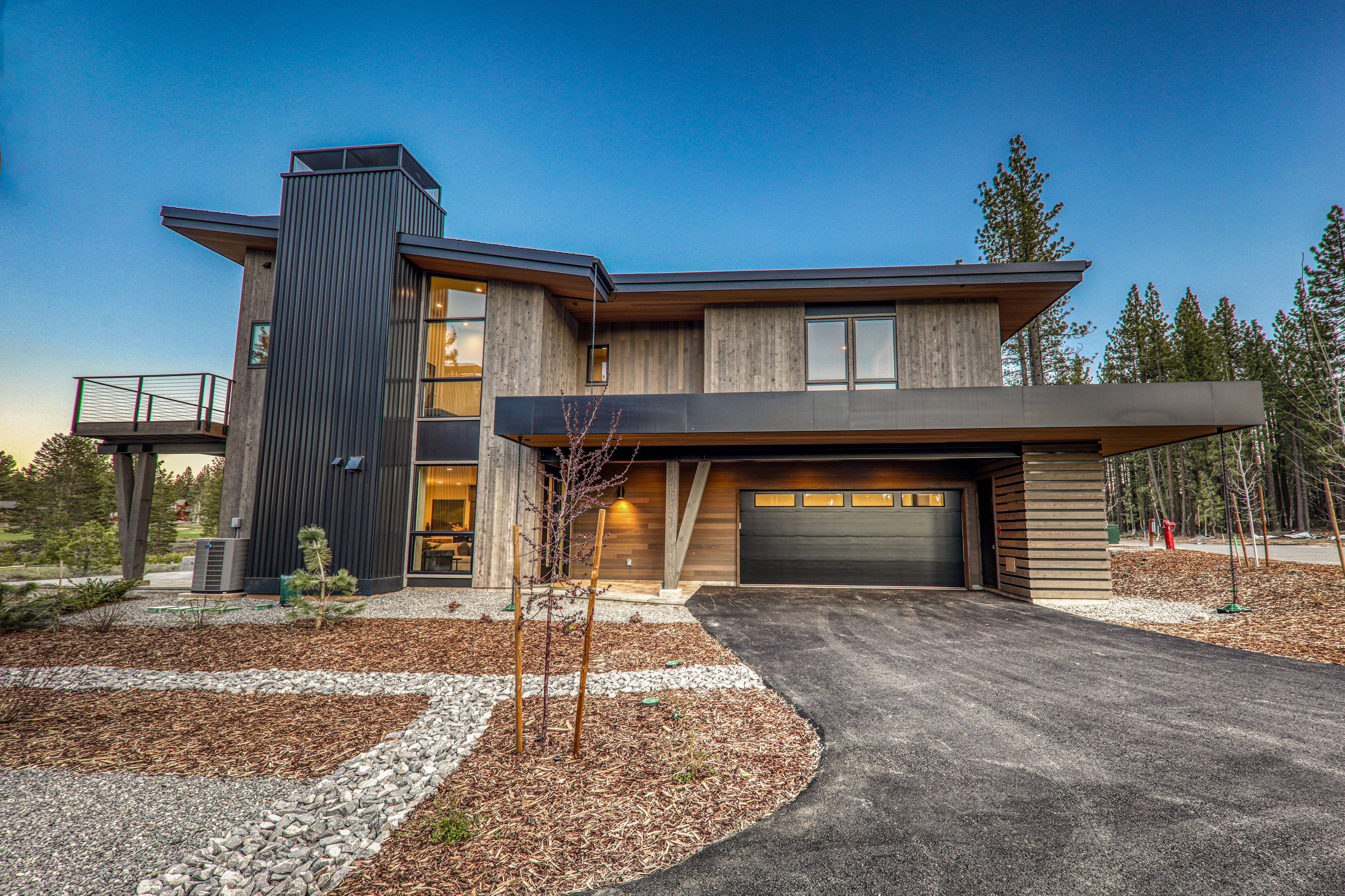 This is a front exterior view of a modern two-story home with a unique architectural design. The house features a combination of wood and metal siding, large windows, and a covered garage. The landscaping includes a mix of gravel, mulch, and young trees, complementing the contemporary style of the property.