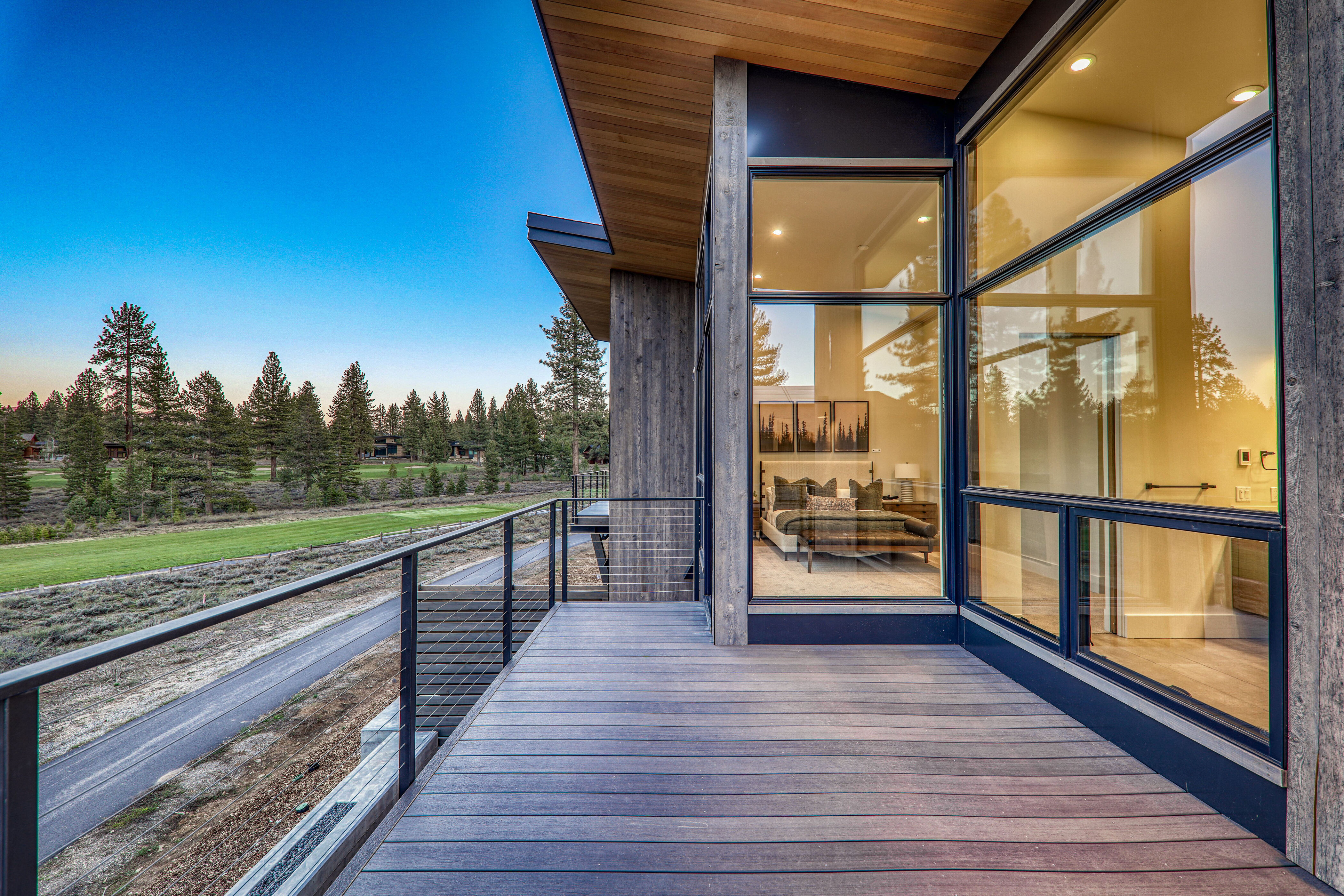 This image showcases a modern home's expansive wooden deck or balcony, offering stunning views of a lush green golf course and surrounding pine trees under a clear blue sky. The deck features sleek metal railings and seamlessly integrates with the home's contemporary architectural design, characterized by large windows that reflect the interior bedroom and bathroom. The perspective is from the deck, looking out towards the landscape, emphasizing the outdoor living space and the connection to nature.