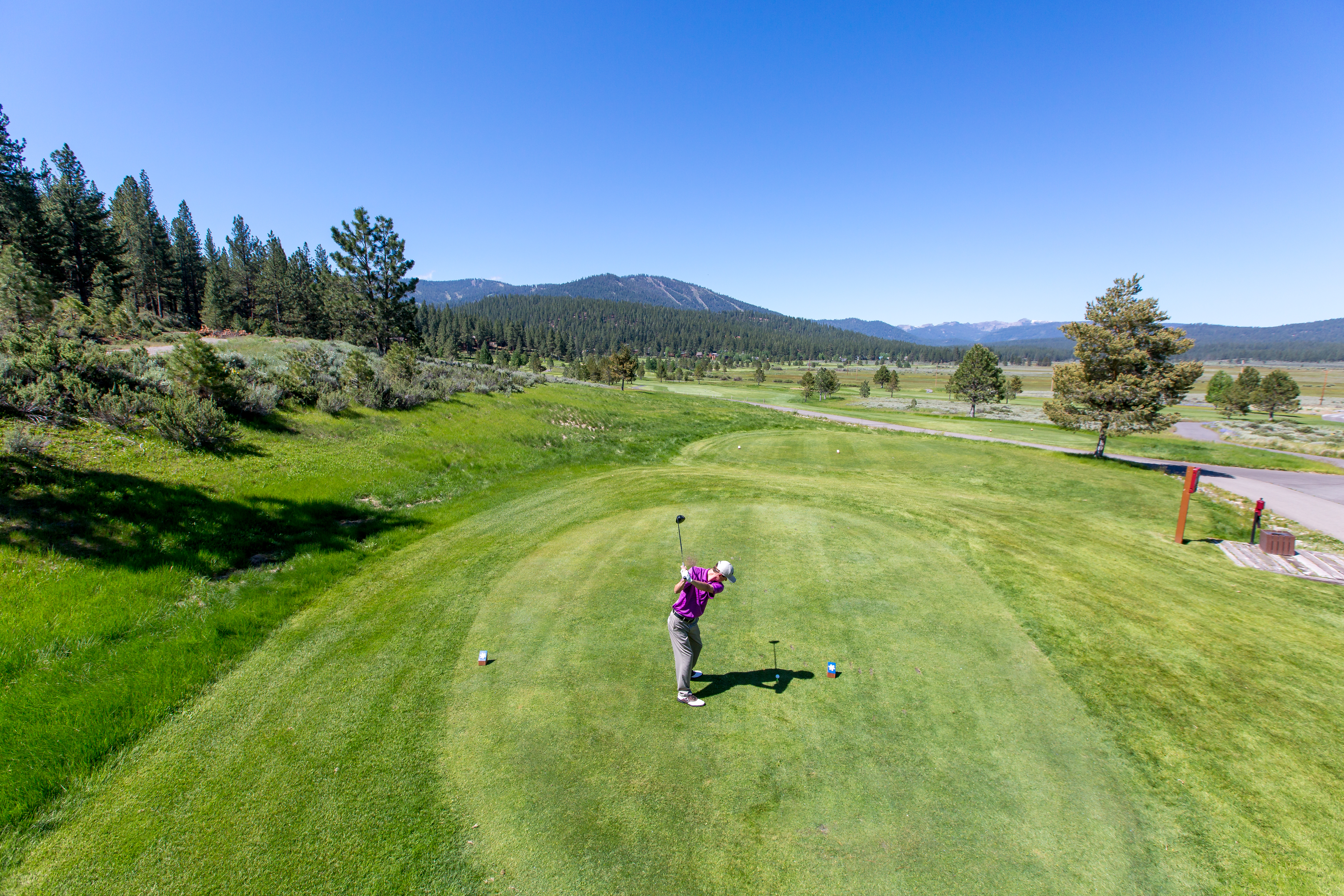 This high-angle aerial shot captures a golfer mid-swing on a pristine, lush green tee box at a scenic golf course. The expansive landscape features a backdrop of dense pine forests and rolling mountains under a clear blue sky, highlighting the property's tranquil and recreational appeal. The perspective emphasizes the vast, well-maintained grounds and the peaceful outdoor environment.