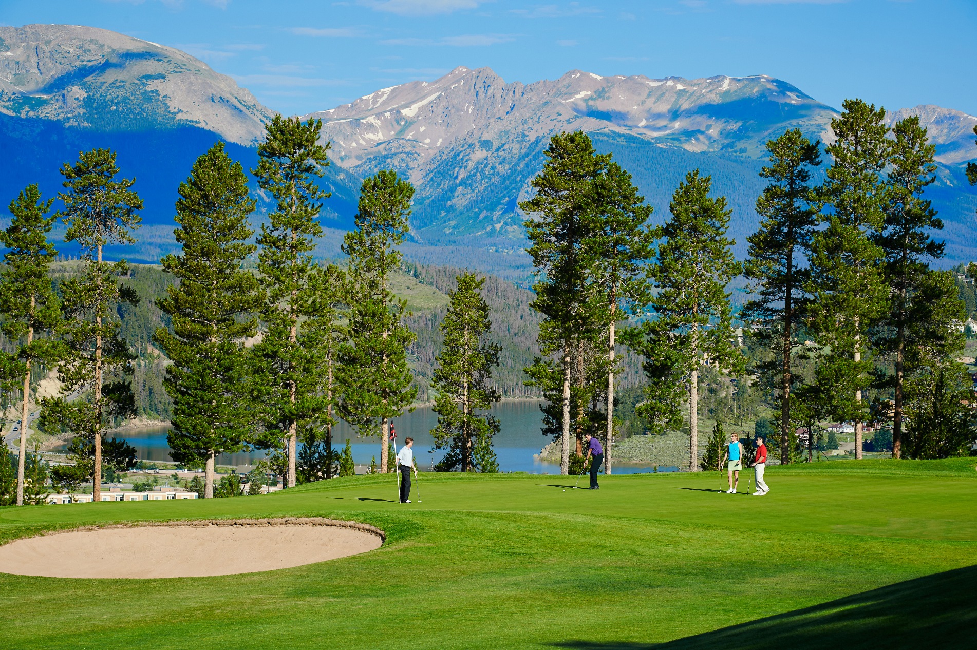 This image showcases a picturesque golf course with a stunning mountain backdrop and a serene lake visible in the mid-ground. Golfers are seen on the green, suggesting an active and recreational lifestyle. The immaculately maintained green, sand trap, and lush trees enhance the aesthetic appeal and highlight the course's amenities.