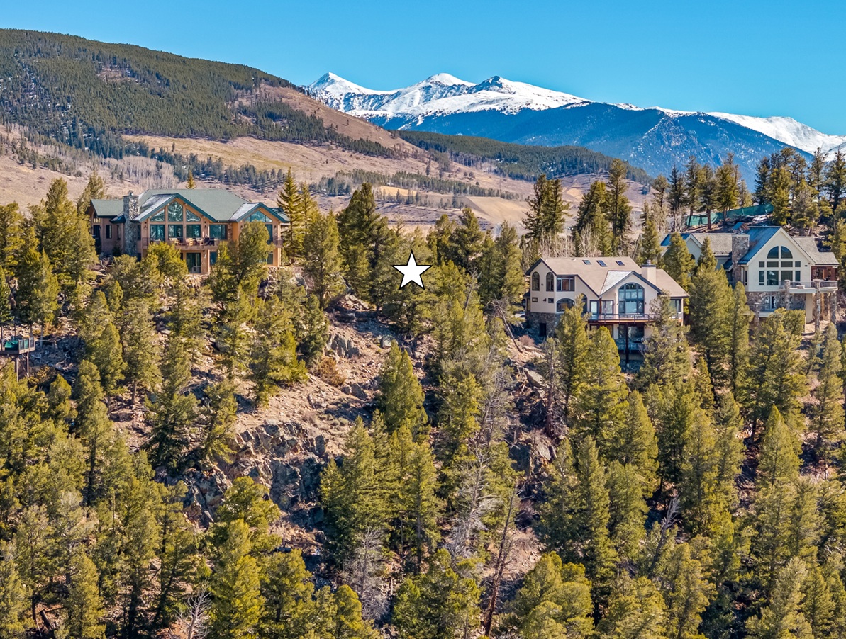 This aerial view showcases an upscale mountain home development nestled amidst lush greenery and towering trees. The large, luxurious homes feature architectural designs that blend seamlessly with the natural landscape. Snow-capped mountains provide a breathtaking backdrop, enhancing the desirability and exclusivity of this real estate offering.