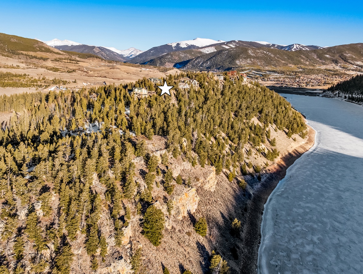 This aerial view showcases a luxurious property nestled on a wooded hillside, potentially overlooking a lake or reservoir, with snow-capped mountains in the background. Several large homes are visible, suggesting a high-end neighborhood. The image emphasizes the property's private and scenic location.