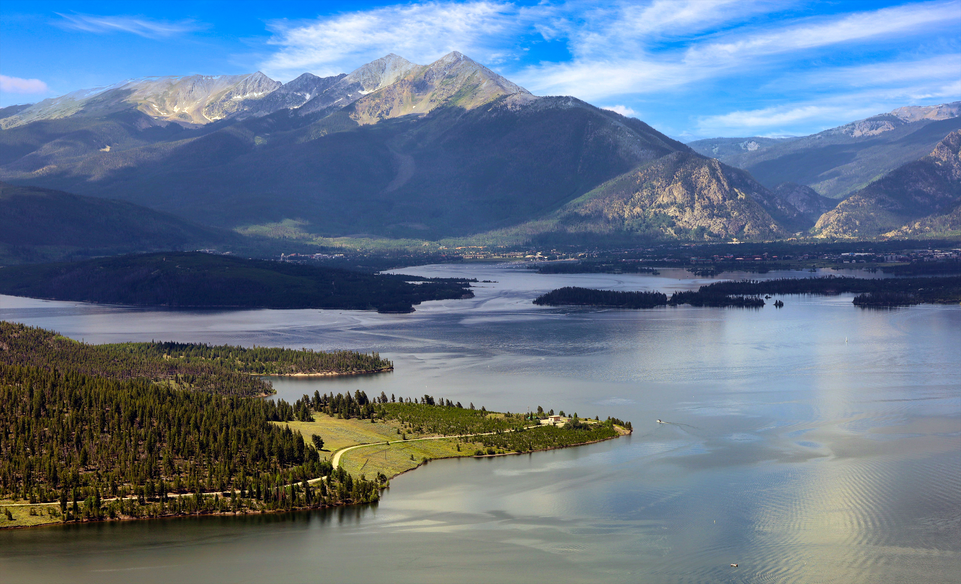 This aerial view showcases a stunning landscape featuring a large, calm lake surrounded by dense forests and majestic mountains in the background. A winding road leads to a building with lawn on a peninsula jutting into the lake providing unique access. The scene is bathed in natural sunlight, projecting a sense of serenity and seclusion, ideal for a lakeside retreat or a nature lover's paradise.