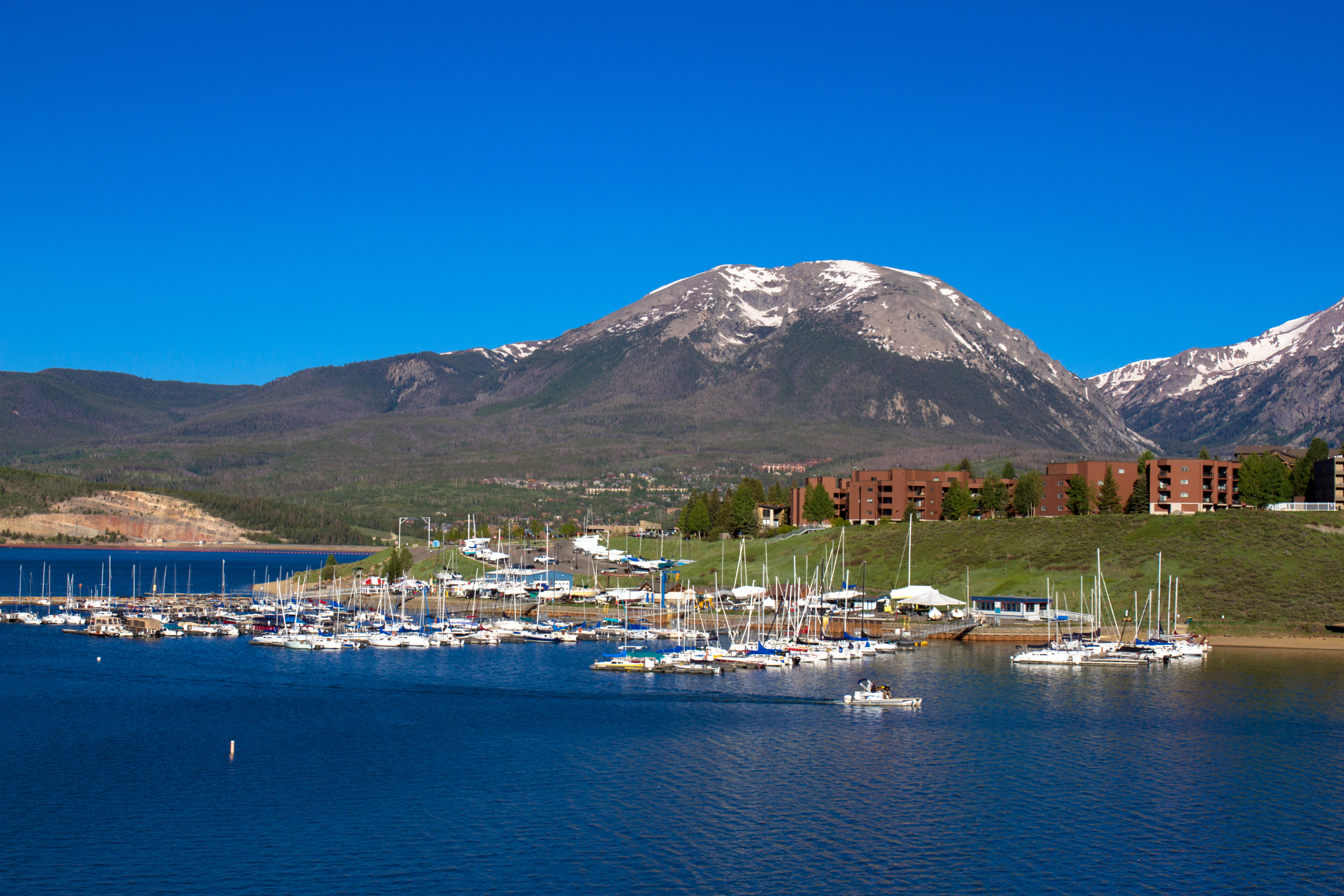 This image showcases a picturesque marina with numerous sailboats and yachts docked. Lush green hillsides and mountains with snow-capped peaks provide a stunning backdrop, suggesting a scenic location. A building complex sits atop the hill, overlooking the marina and adding to the appeal of this waterfront community amenity.