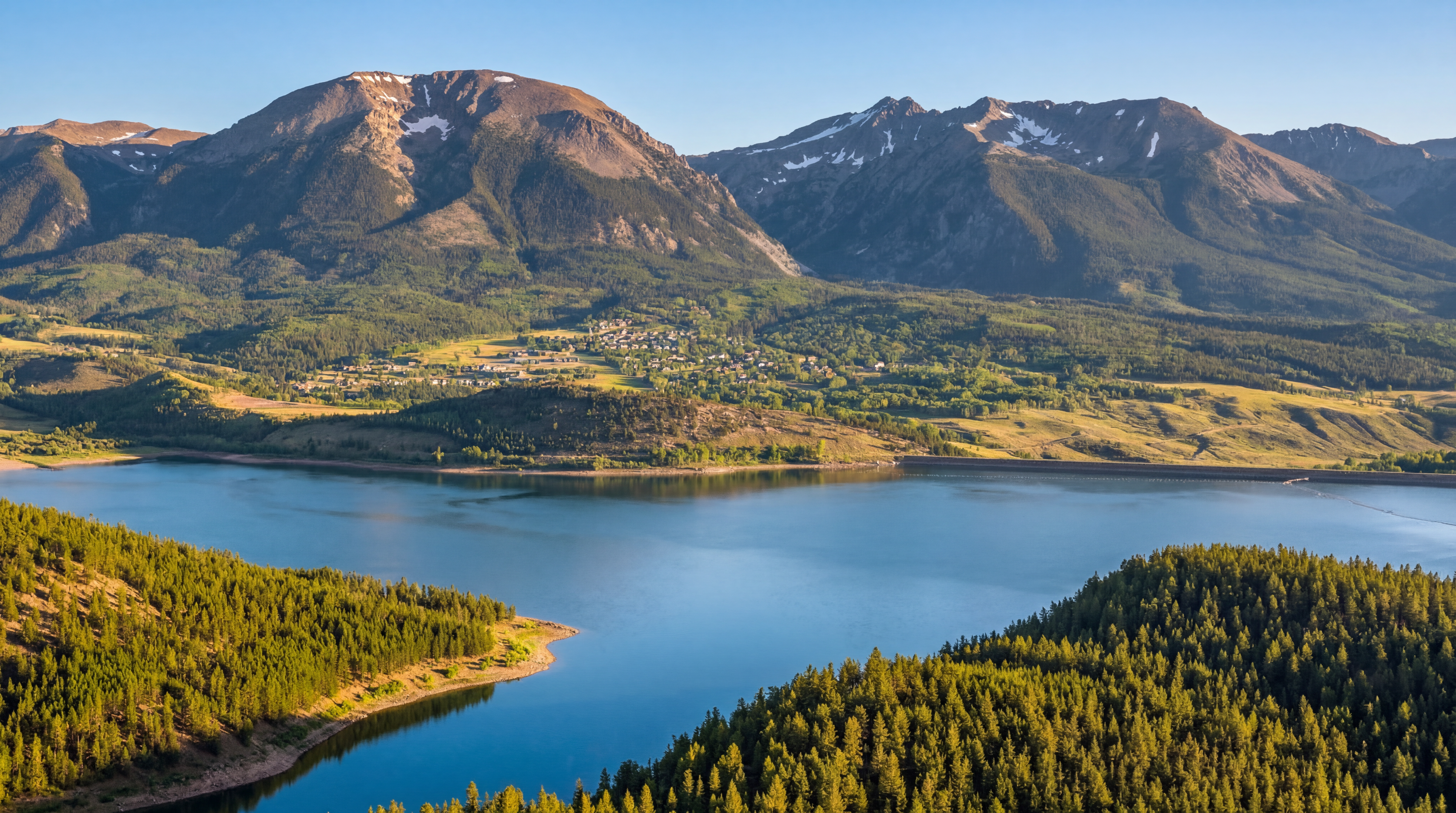 This aerial view captures a stunning landscape featuring a serene lake surrounded by lush green forests and towering mountains. A small town nestled in-between the mountains adds a charming element to the scene. The clear blue sky enhances the peaceful and scenic beauty of the location, ideal for highlighting the natural amenities near potential real estate.