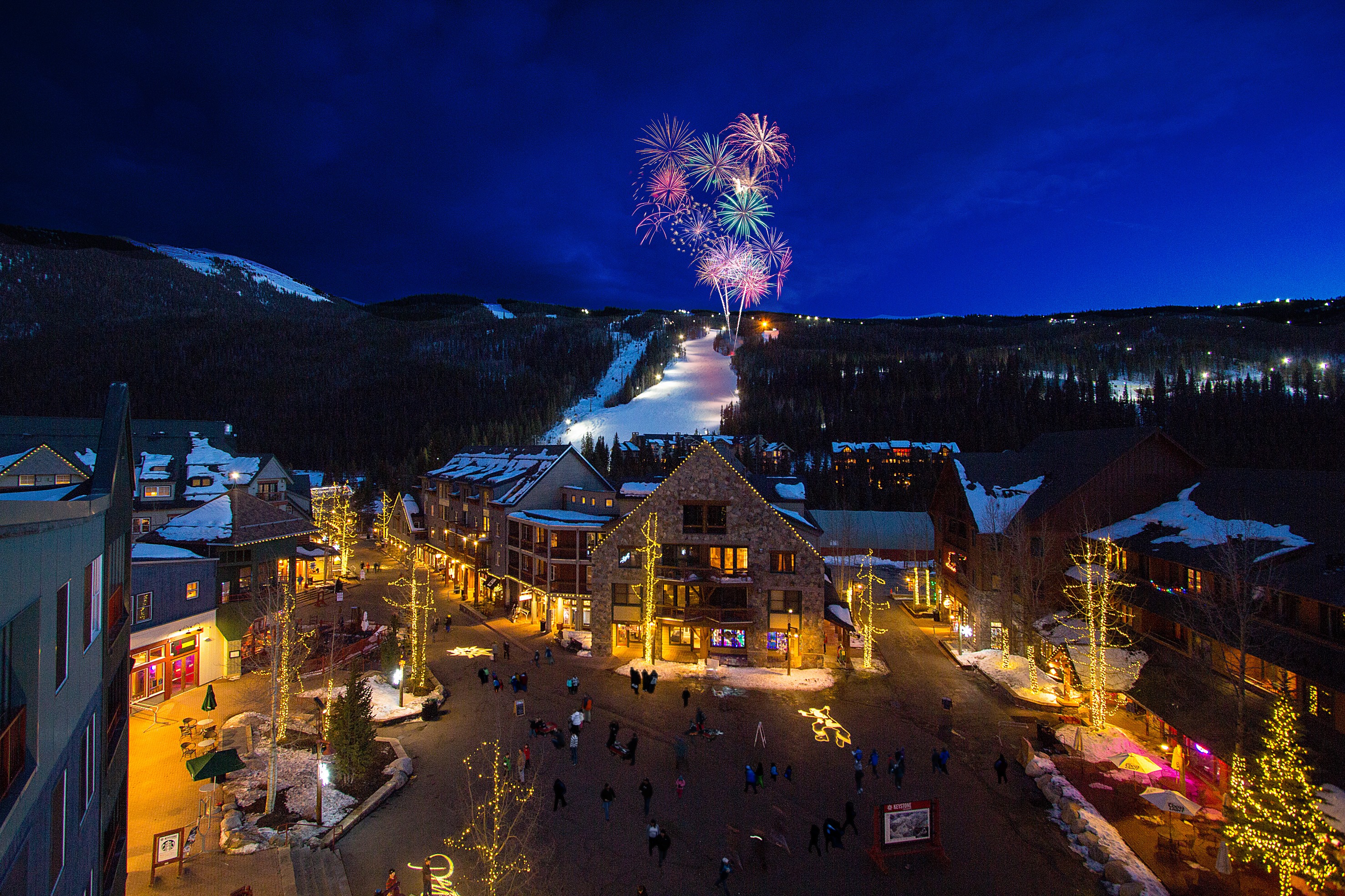 This aerial image captures a lively town square at night, likely a resort or ski village, where fireworks are illuminating the sky. The buildings are warmly lit and decorated, hinting at retail shops and restaurants, while a multitude of people are gathered below, suggesting a festive atmosphere. This image would be ideal for a property within walking distance to the town square.