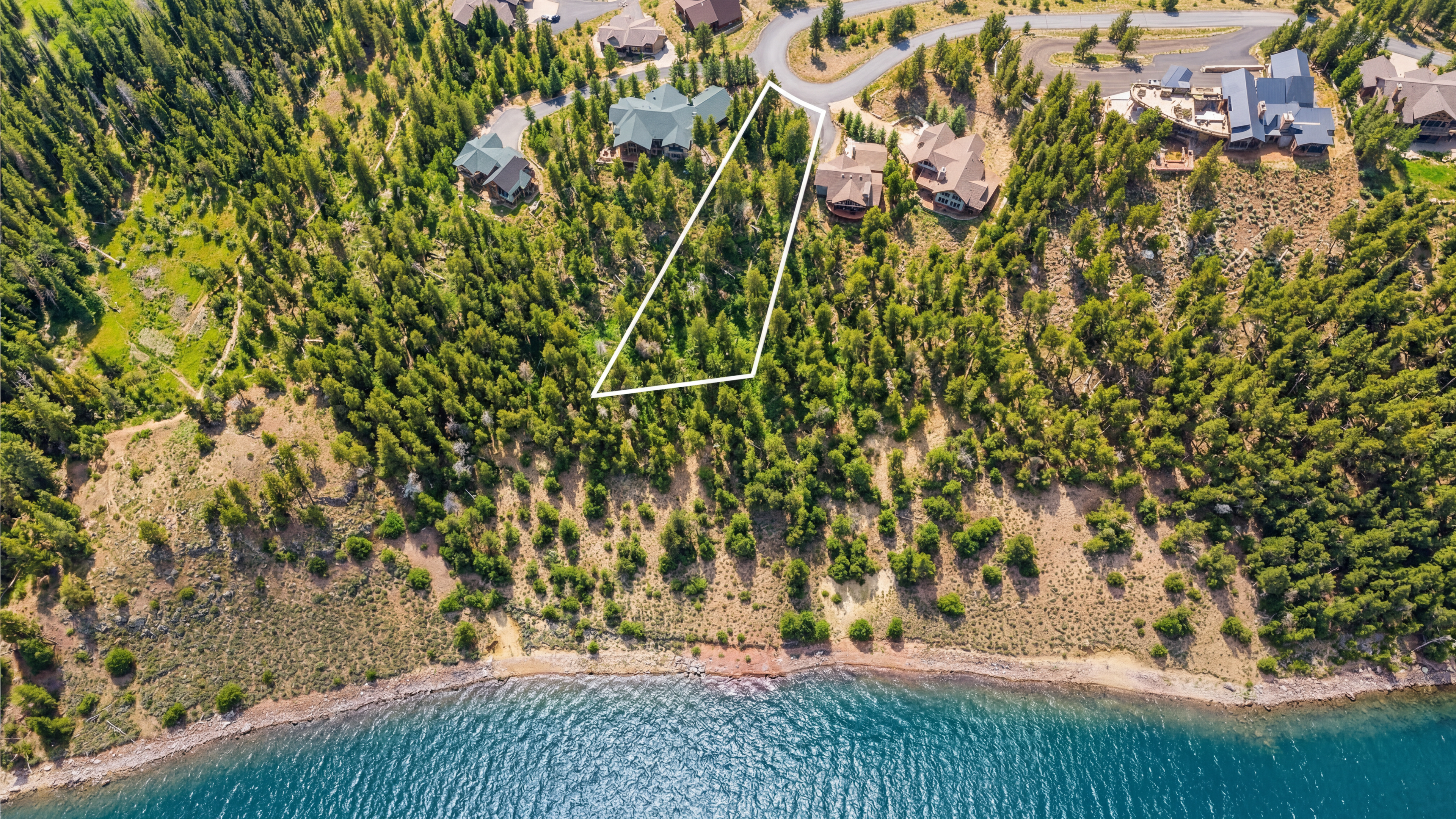 This aerial view showcases a beautiful piece of real estate nestled between lush greenery and a serene lake. A highlighted land plot presents a unique opportunity for development, complemented by nearby luxury homes. The landscape offers both privacy and scenic views, making it an ideal setting for a custom-built residence.