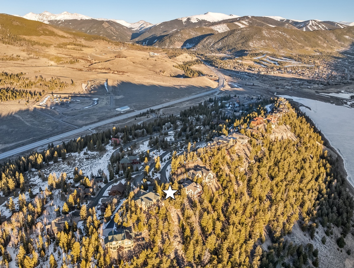 This aerial view showcases a neighborhood nestled on a mountainside, presenting a stunning landscape. Several homes stand out, interspersed among trees and leading up to the property marked with a white star. In the background, snow-capped mountain peaks add to the scenic backdrop.