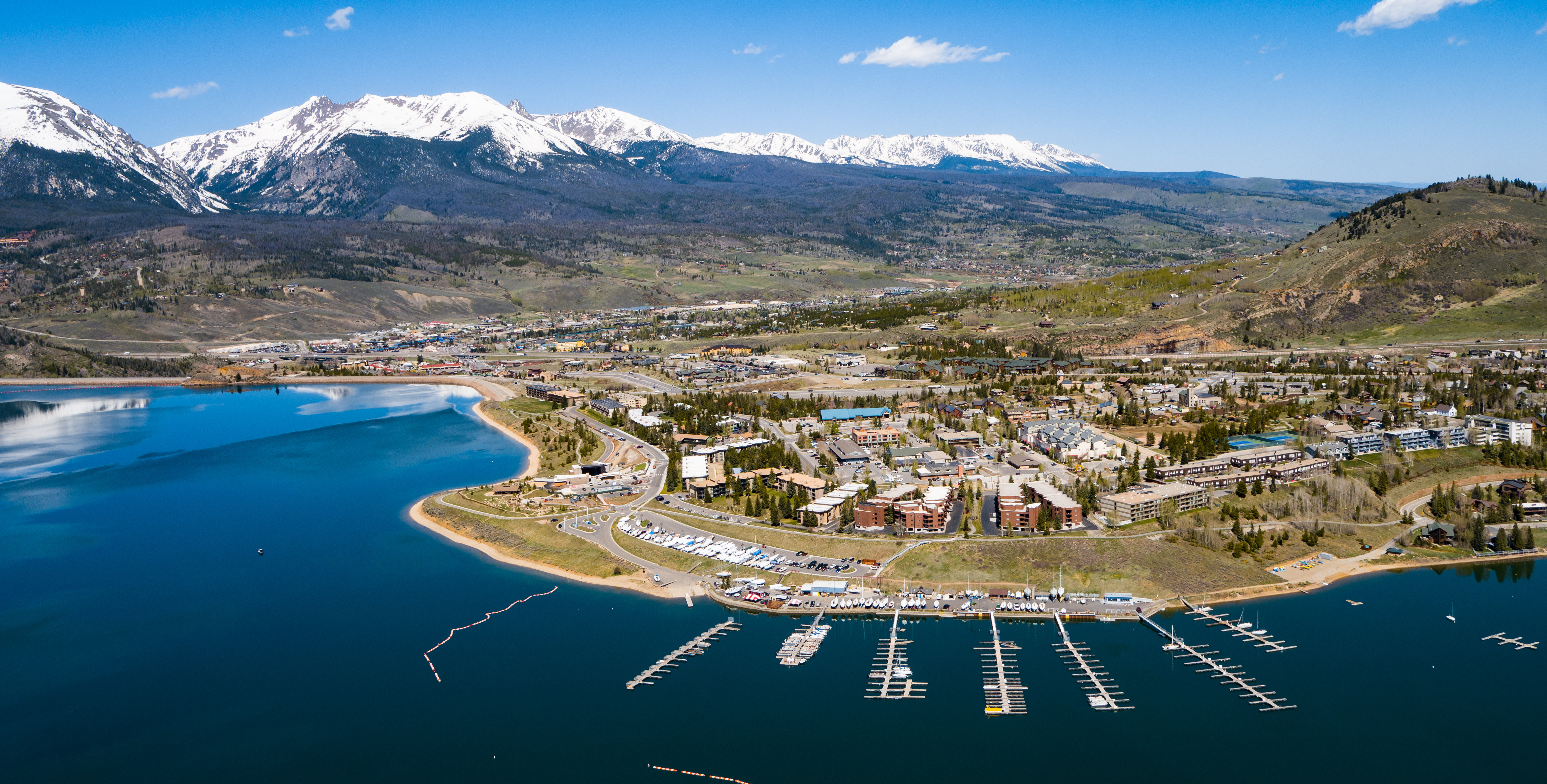 This aerial view showcases a lakeside community nestled against a backdrop of majestic snow-capped mountains and lush greenery. The marina fronting Dillon Reservoir features numerous boats and docks, creating a picturesque waterfront. The neighborhood appears well-maintained with a variety of housing styles, indicating a desirable location with scenic amenities.