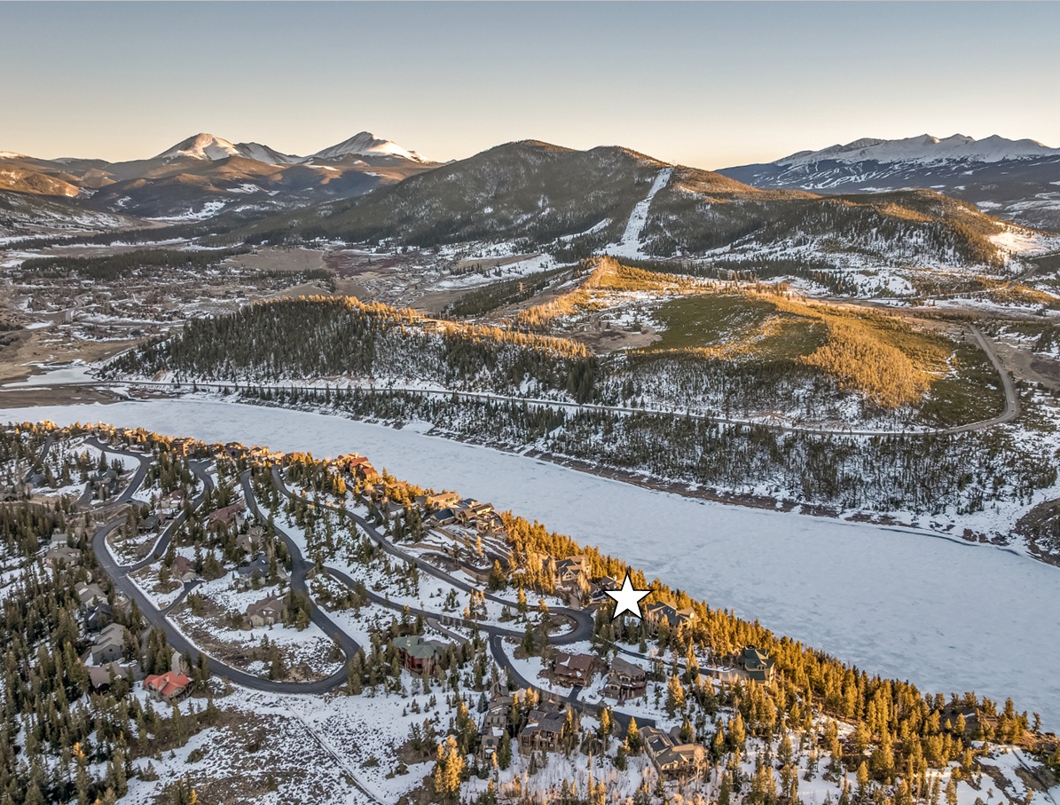 This is an aerial view of a residential area nestled in a snowy mountain landscape. The scene features a winding road leading through clusters of houses, with a frozen lake stretching out nearby. Mountains rise in the background, creating a scenic and serene setting.