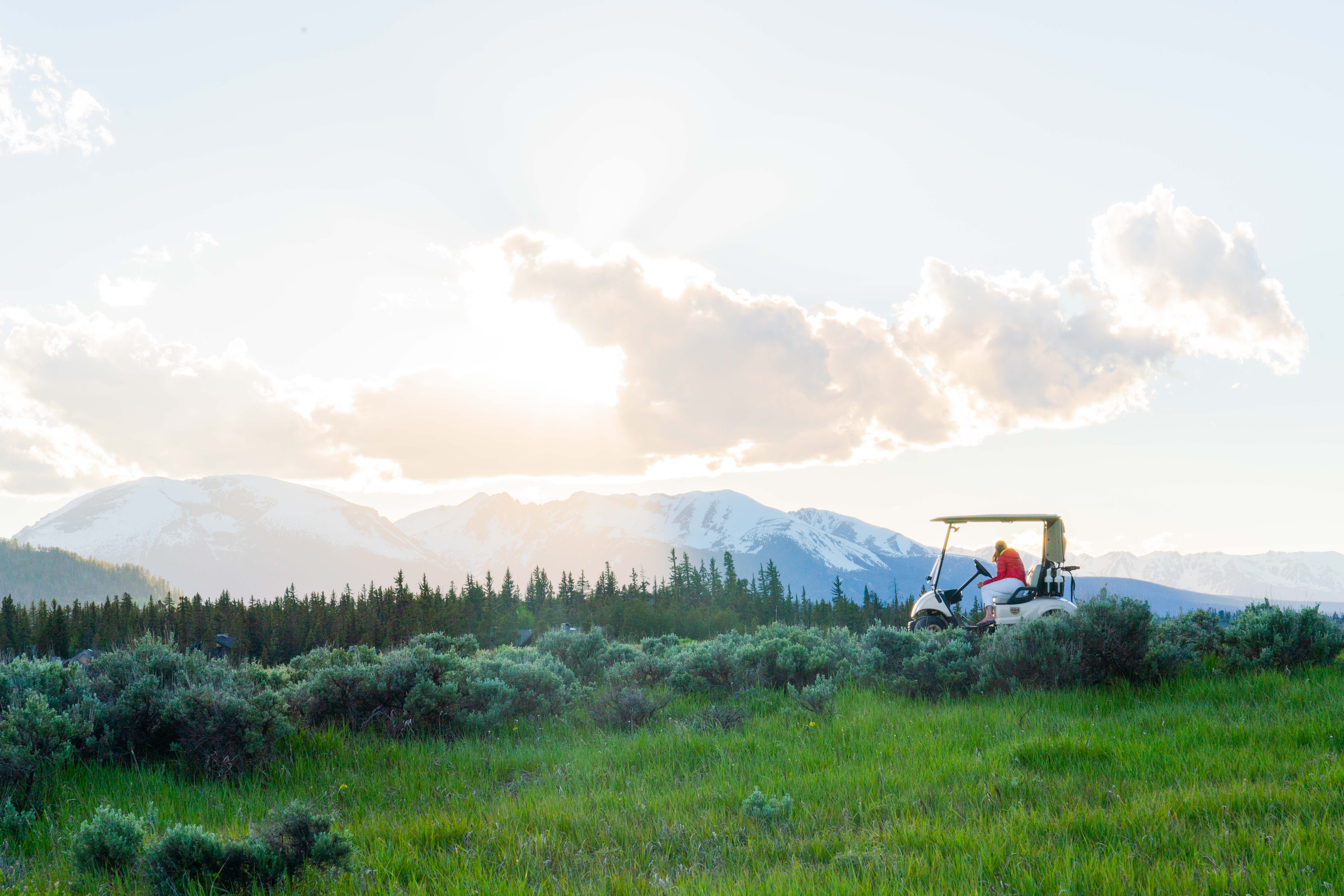 This image showcases a vibrant outdoor scene with green grass and shrubbery in the foreground, transitioning into a lush landscape. A golf cart is parked amidst the scenery, and a mountain range with snow-capped peaks provides a scenic backdrop. The image evokes a sense of tranquility and outdoor recreation, highlighting the property's connection to nature.