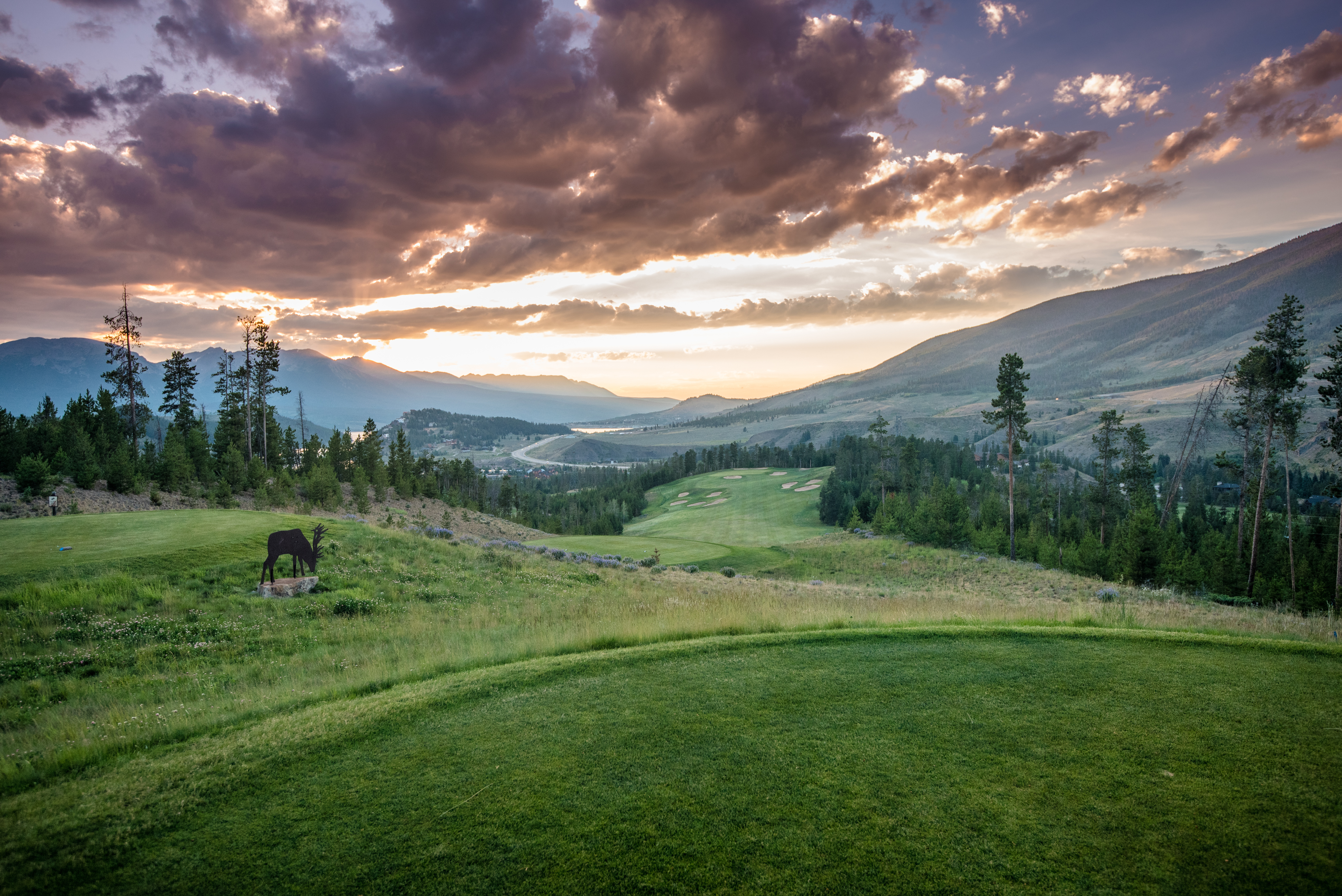 This image showcases a scenic golf course set against a backdrop of mountains and a vibrant sunset. The manicured green of the tee box transitions to the fairway and includes a decorative elk statue. The view highlights the recreational opportunity and natural beauty associated with this community.