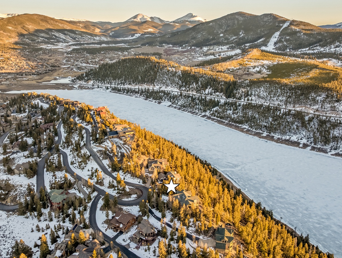 This aerial view showcases a high-end residential area nestled near a frozen lake and mountains. The homes feature a variety of architectural styles, with winding roads connecting the properties. The landscape is a blend of snow-covered ground and evergreen trees, presenting a serene yet luxurious living environment, and the property marked by the star has scenic views.