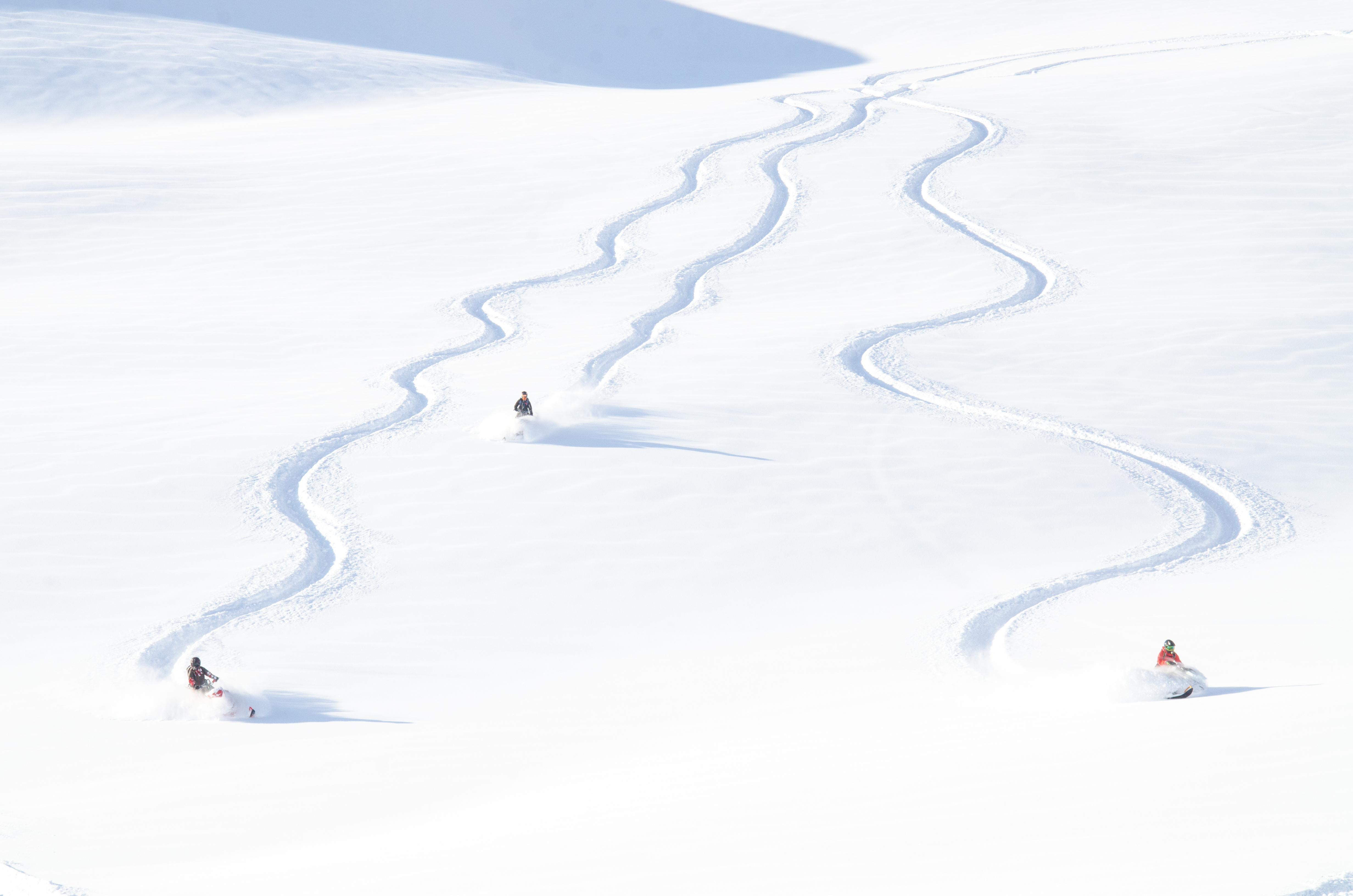 The image captures a vast, snow-covered landscape with multiple snowmobile tracks creating dynamic lines across the scene. Several individuals on snowmobiles add a sense of motion and adventure. The bright, expansive white field suggests a recreational area perfect for winter sports, which may be of interest for properties near such landscapes.
