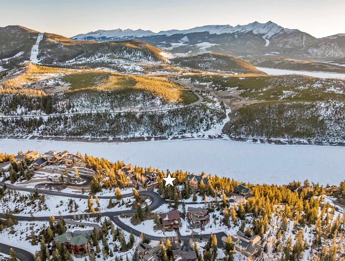 This is an aerial view of a luxury residential area near a frozen lake or reservoir. The homes are large and appear well-maintained, situated among snow-covered pine trees. The curved roads suggest a secluded and upscale neighborhood with a stunning backdrop of snow-capped mountains.