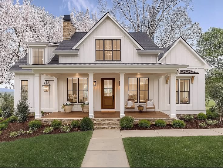 This charming modern farmhouse features white board-and-batten siding, a prominent gable roof, and a welcoming covered front porch. The exterior is accented by a stone chimney, black-framed windows, and a rich wood front door, set against a backdrop of blooming cherry blossom trees. The neatly landscaped lawn and paved walkway create excellent curb appeal, embodying a classic yet contemporary aesthetic.