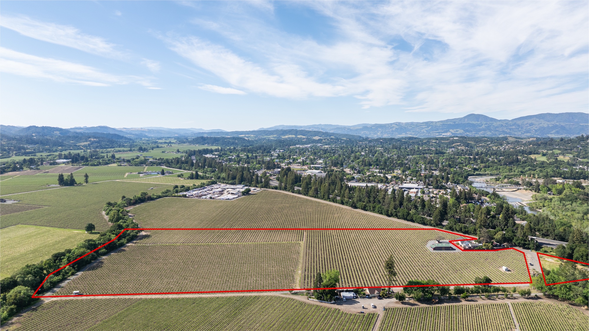 This stunning aerial view highlights a sprawling vineyard property delineated by a clear red outline. The estate features vast rows of grapevines, established structures, and scenic surroundings, suggesting a prime location for agriculture or estate development. The elevated perspective captures the expansive nature of the land and its integration into the surrounding landscape.