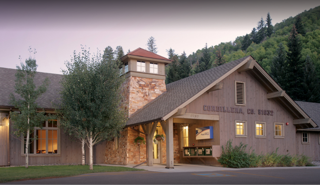 This image captures the front exterior of a rustic, mountain-style clubhouse or community center, characterized by its stone tower and wood-paneled siding. The structure is nestled against a lush, forested hillside, creating a serene and inviting atmosphere. The perspective is a wide, eye-level shot that highlights the building's architectural charm and its integration into the natural landscape.