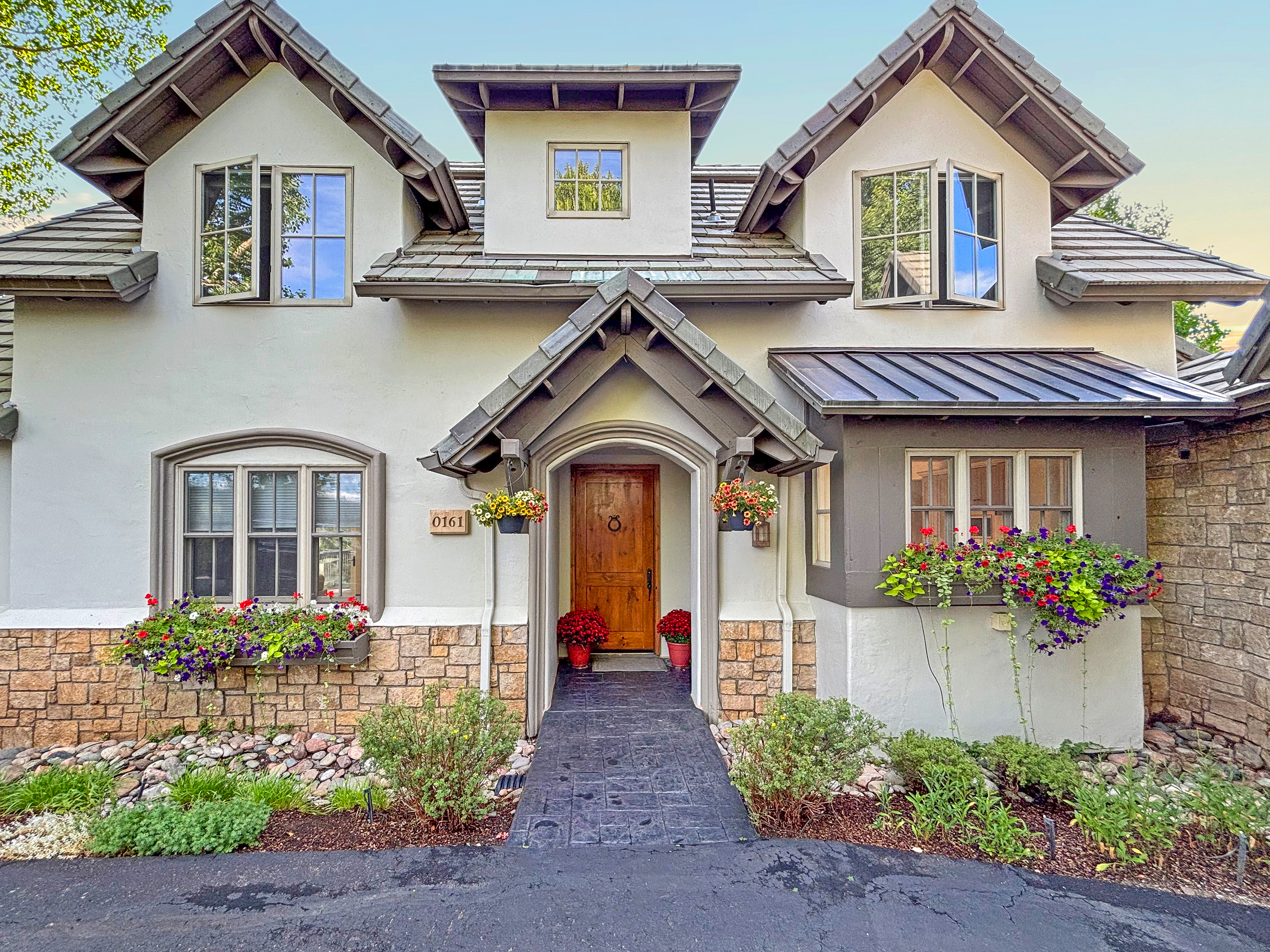 This charming, multi-gabled home features a blend of stucco and stone masonry, creating a warm and inviting cottage-style aesthetic. The front entrance is highlighted by a covered porch with a wooden door, flanked by vibrant flower boxes and potted plants that add a splash of color to the facade. The perspective is a straight-on, eye-level shot that captures the symmetrical architectural details and the well-maintained landscaping.