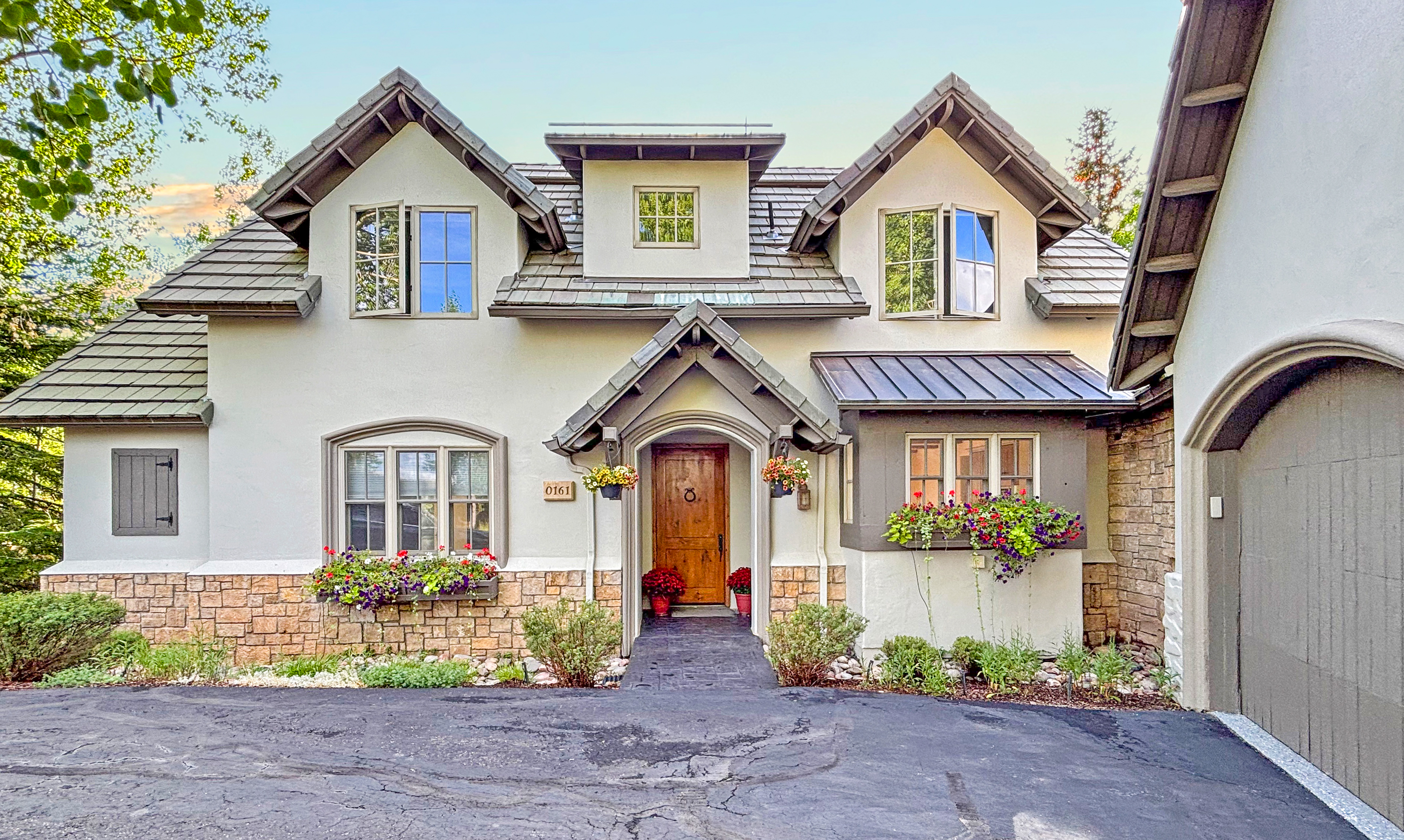 This charming, storybook-style home features a symmetrical facade with a prominent central entryway, dual gabled dormers, and a blend of stucco and stone masonry. The exterior is adorned with vibrant window boxes overflowing with colorful flowers, creating a welcoming and picturesque curb appeal. The perspective is a straight-on, eye-level shot that captures the full architectural character and inviting atmosphere of the residence.