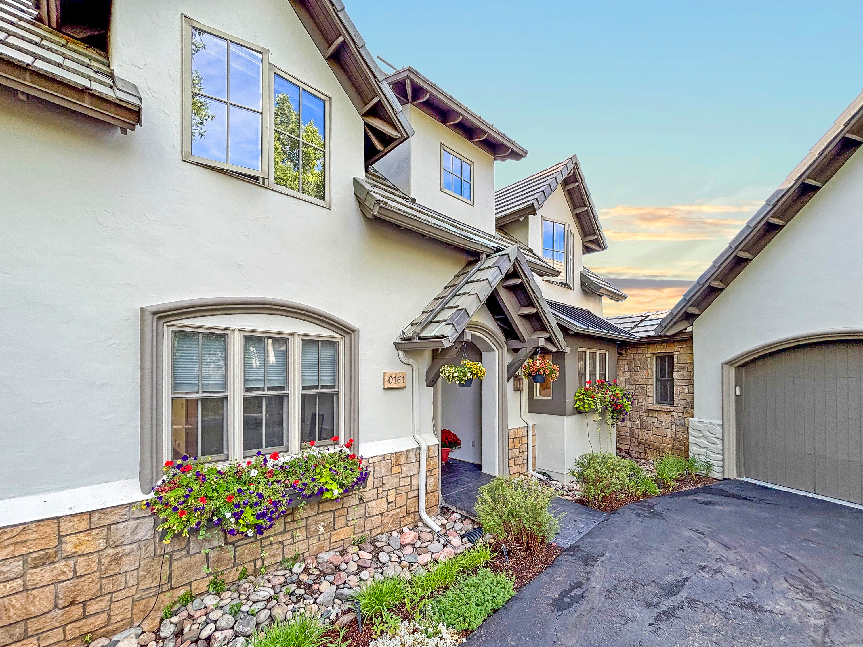 This charming home features a blend of stucco and stone siding, accented by a gabled entryway and a prominent window box filled with vibrant flowers. The perspective captures the inviting front entrance and a portion of the attached garage, set against a clear sky. The overall aesthetic is warm and welcoming, highlighting the home's curb appeal and well-maintained landscaping.