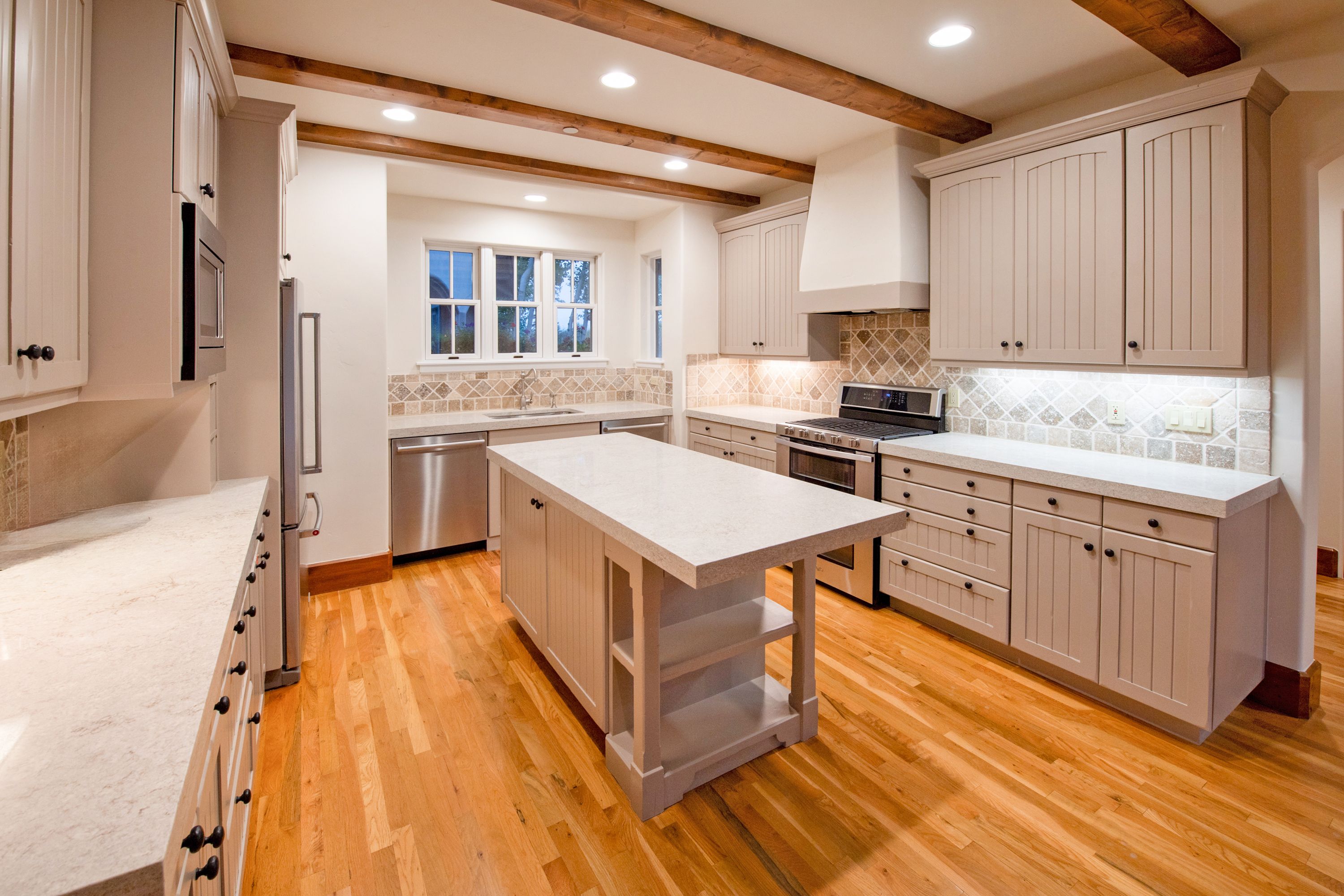 This bright and inviting kitchen features a classic farmhouse aesthetic with light-toned cabinetry, a central island with open shelving, and warm hardwood flooring. The space is accented by rustic wooden ceiling beams, a tiled backsplash, and high-end stainless steel appliances, creating a functional yet cozy atmosphere. The wide-angle perspective captures the entire layout, highlighting the seamless flow between the prep areas and the central island.