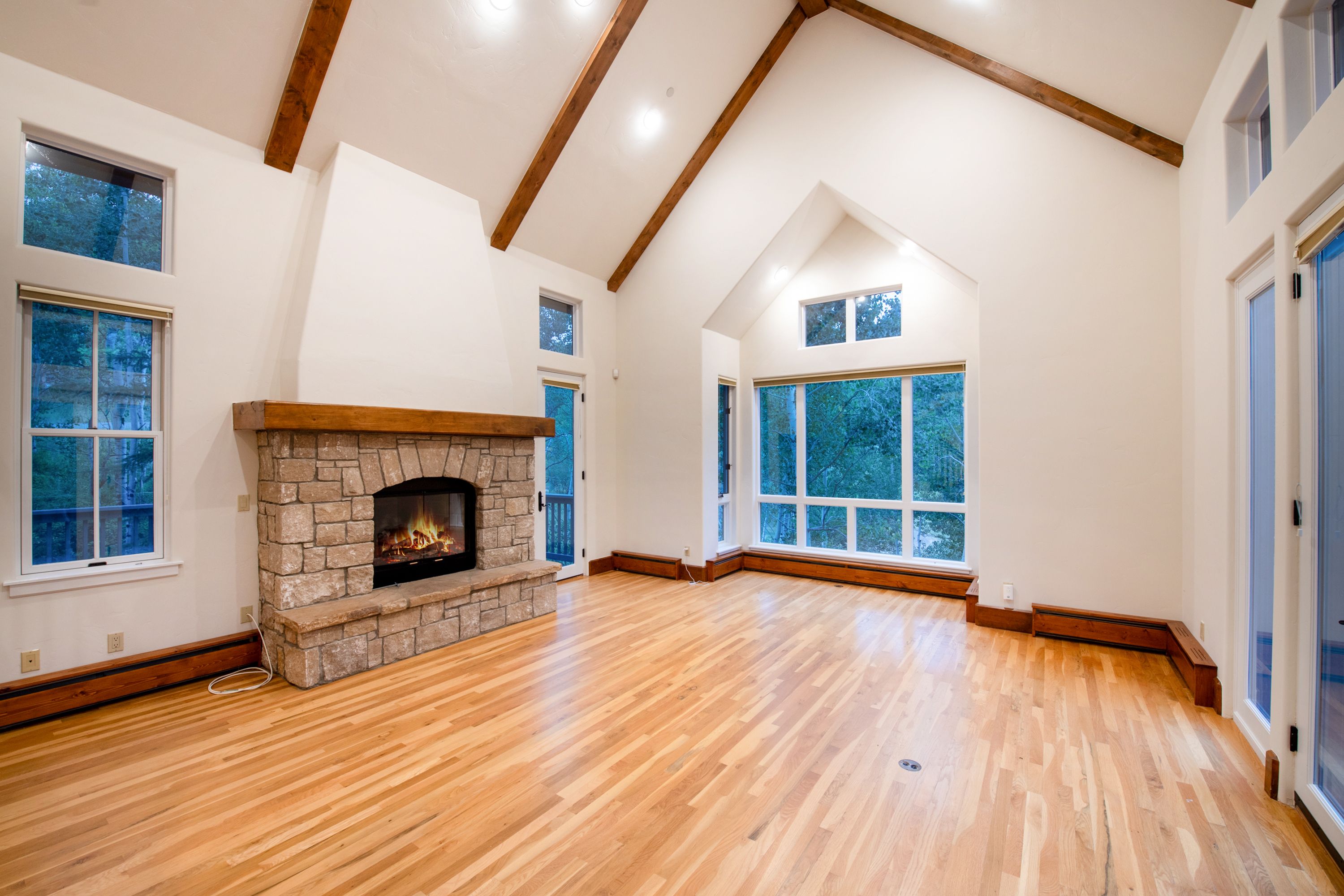 This spacious living room features a dramatic vaulted ceiling with exposed wooden beams and a striking floor-to-ceiling stone fireplace as its centerpiece. Large windows flood the room with natural light, offering serene views of the surrounding trees, while the warm hardwood flooring adds a cozy, inviting touch. The open-concept layout and clean white walls create an airy, sophisticated atmosphere perfect for comfortable living.