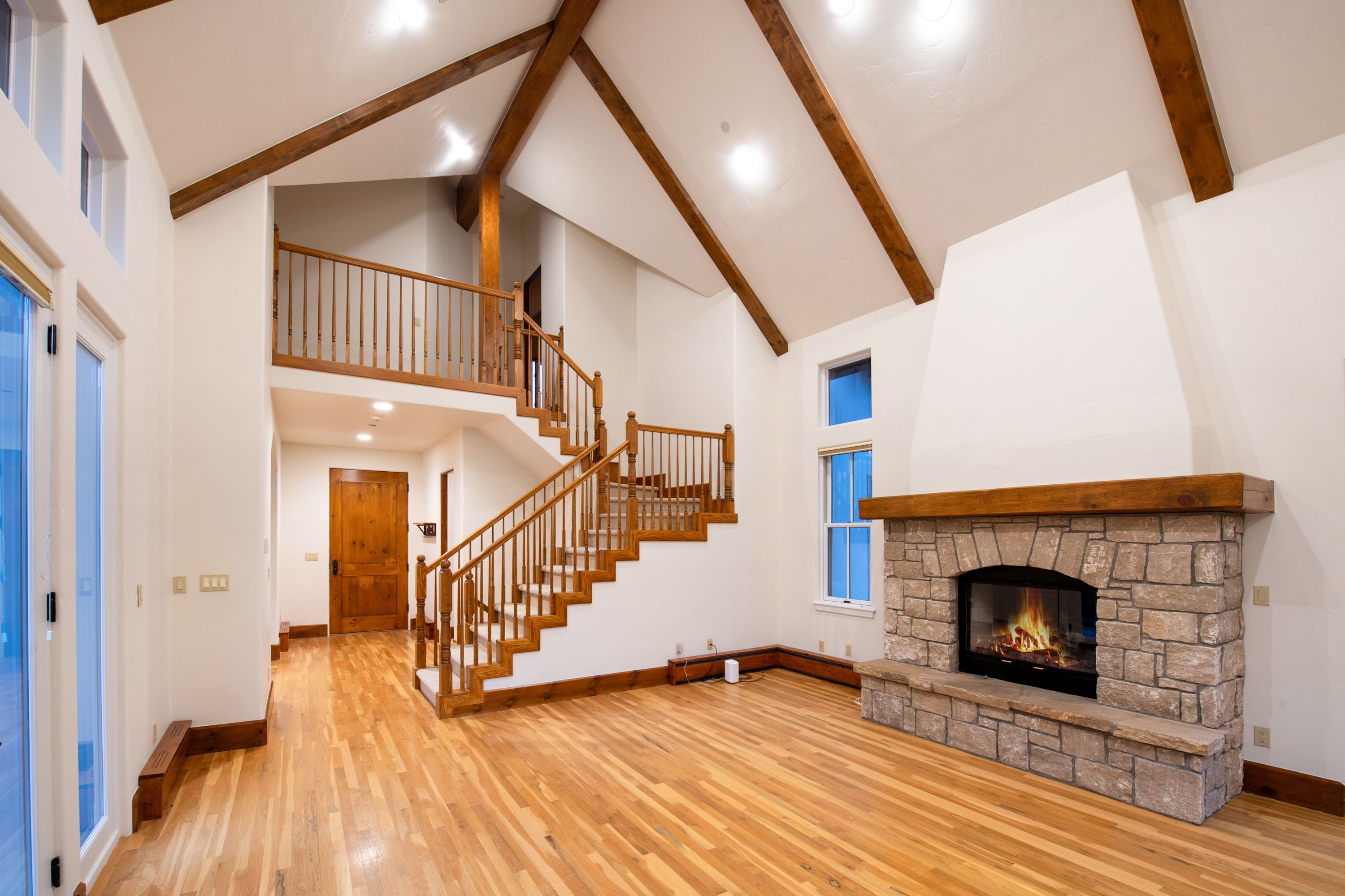 This spacious living room features a dramatic vaulted ceiling with exposed wooden beams and a striking stone fireplace as the focal point. A wooden staircase with a matching railing leads to an upper landing, creating an open and airy architectural flow. The warm hardwood floors and natural wood accents throughout the space provide a cozy, rustic-modern aesthetic.