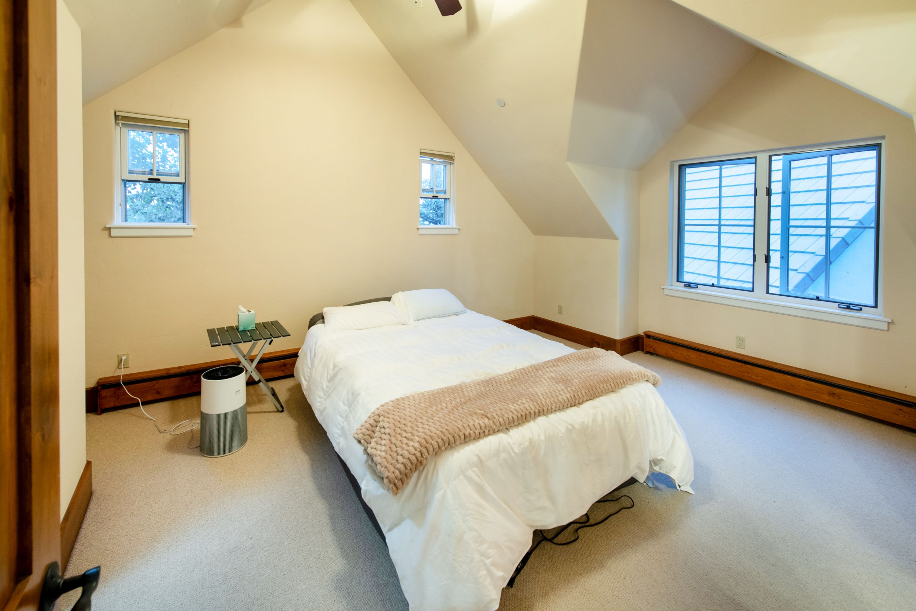 This cozy, carpeted bedroom features a vaulted ceiling and a neutral color palette, creating a bright and airy atmosphere. The room is furnished with a comfortable bed topped with a textured throw blanket, a small folding side table, and an air purifier, while multiple windows allow for ample natural light. The space is simple and functional, offering a peaceful retreat under the sloped roofline.