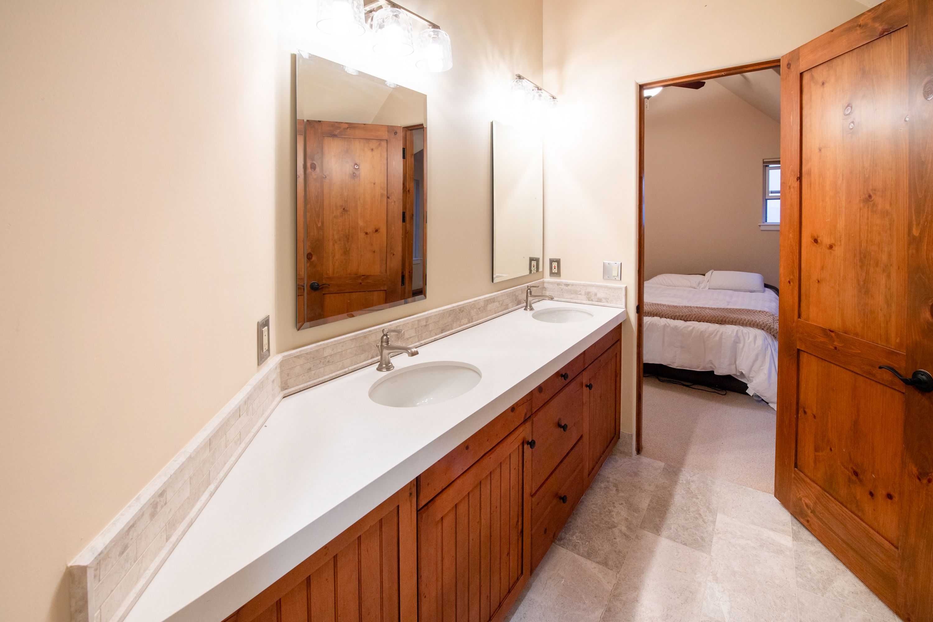 This primary bathroom features a long, dual-sink vanity with a white countertop, stone backsplash, and warm-toned wooden cabinetry. The space is illuminated by overhead vanity lighting and includes two mirrors, with a view leading directly into a cozy, carpeted bedroom. The overall aesthetic is rustic and inviting, blending natural wood textures with clean, functional design.