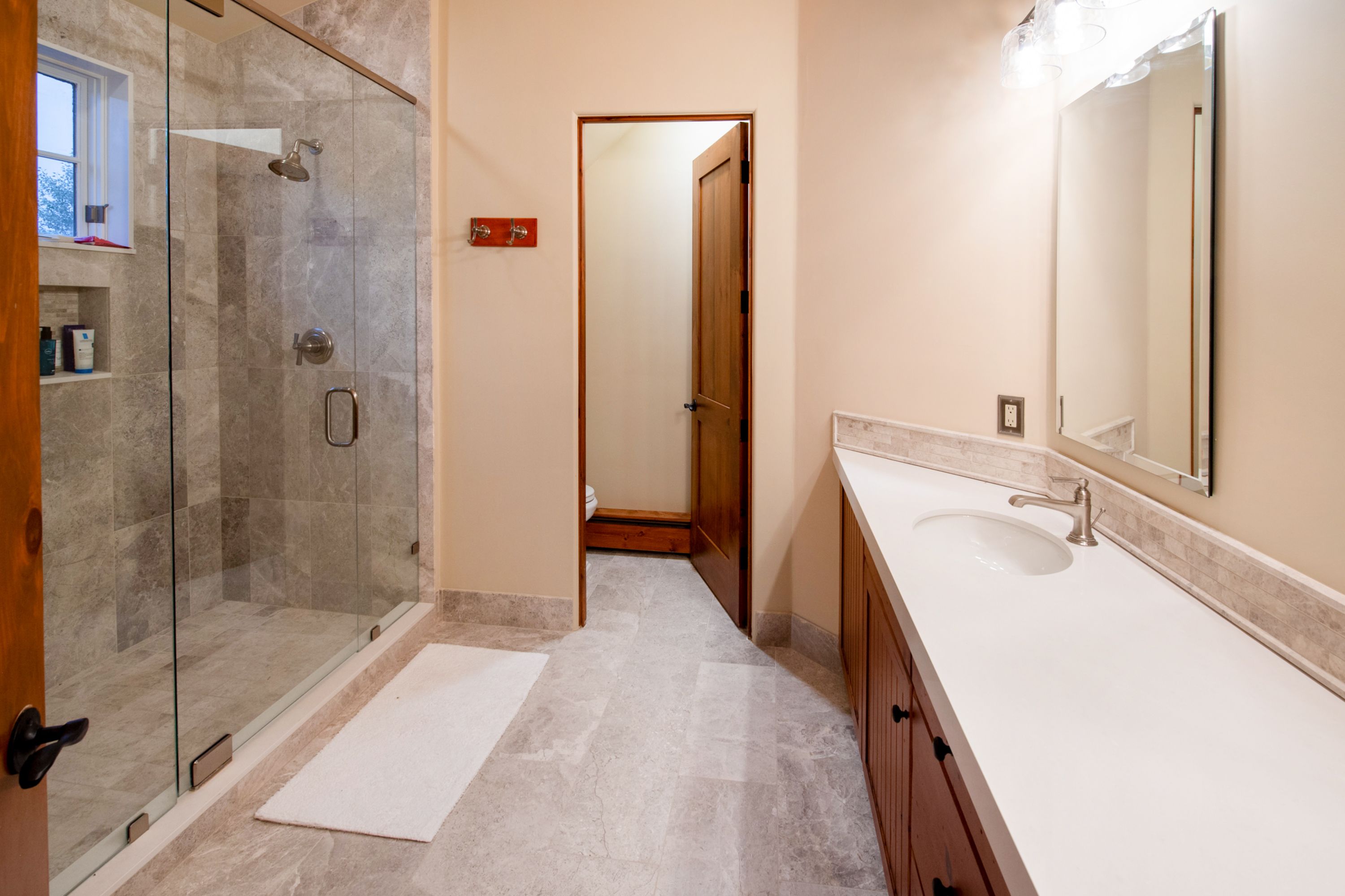 This well-appointed bathroom features a spacious walk-in glass shower with grey stone tile and a separate water closet area. A long vanity with a white countertop and warm wood cabinetry provides ample storage, complemented by a large wall-mounted mirror. The neutral color palette and clean lines create a bright, functional, and inviting space.