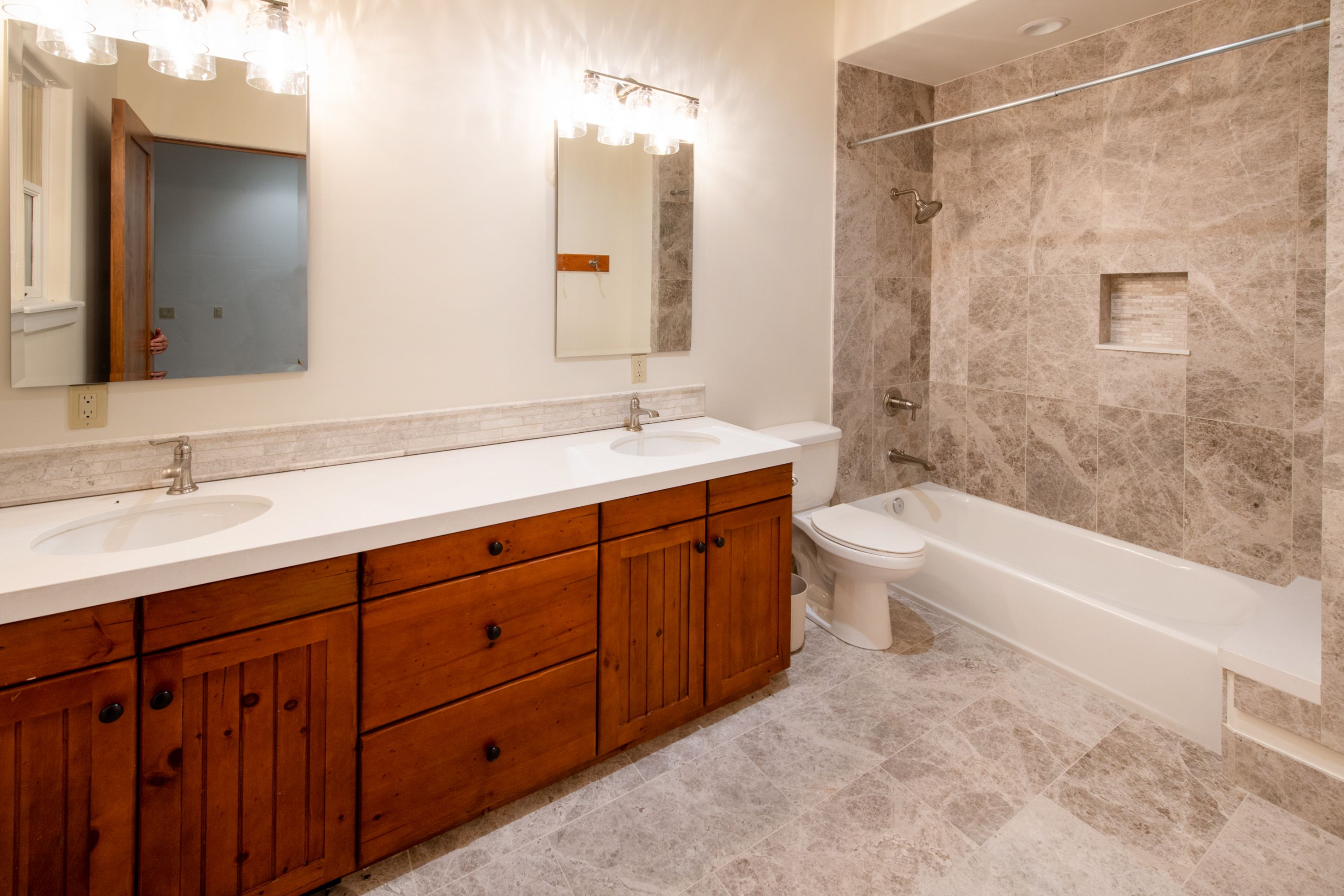 This spacious primary bathroom features a dual-sink vanity with warm wood cabinetry and a clean white countertop, complemented by two mirrors and modern lighting fixtures. The room is finished with elegant, large-format marble-look tile flooring and a matching tiled shower-tub combination. The overall aesthetic blends rustic wood elements with a sophisticated, neutral stone palette for a clean and inviting atmosphere.