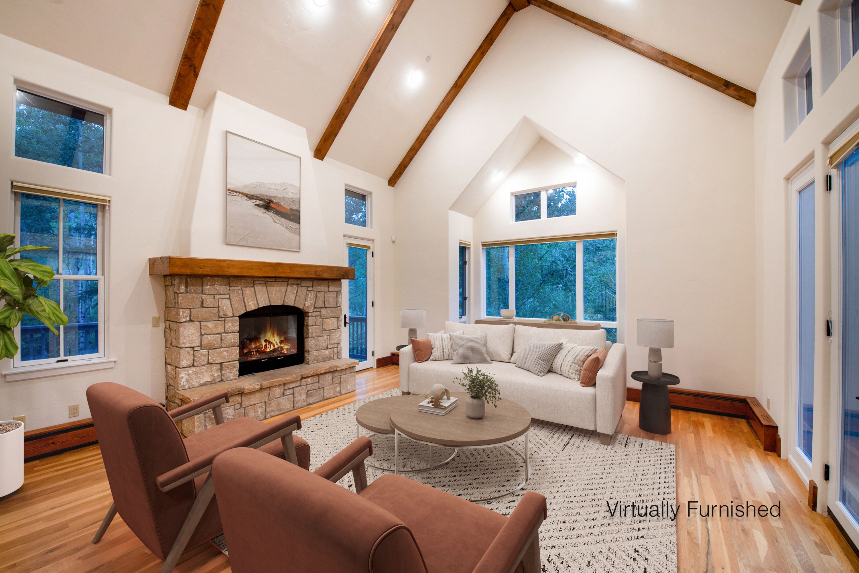 This spacious living room features a dramatic vaulted ceiling with exposed wooden beams and a striking stone fireplace as its focal point. The room is bathed in natural light from large windows and glass doors, creating an airy, inviting atmosphere that blends rustic charm with modern comfort. The space is tastefully furnished with a neutral sofa, two accent chairs, and a layered coffee table set, all resting on a textured area rug.