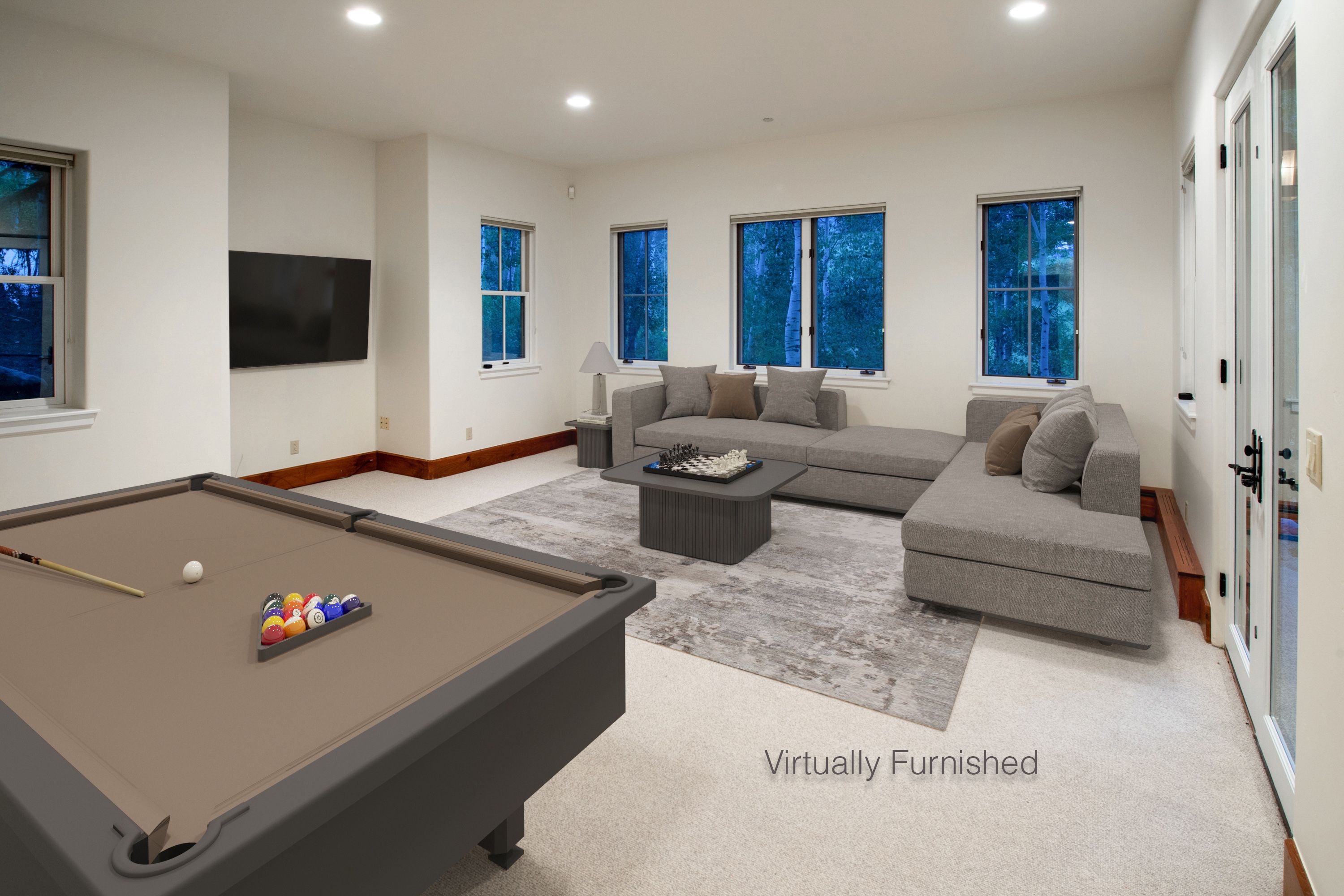 This spacious, virtually furnished living area features a contemporary gray sectional sofa arranged around a modern coffee table, creating a comfortable lounge space. A pool table occupies the foreground, while a wall-mounted television and large windows overlooking a wooded area add to the room's entertainment appeal. The neutral color palette and clean lines provide a versatile and inviting atmosphere suitable for a game room or casual living space.