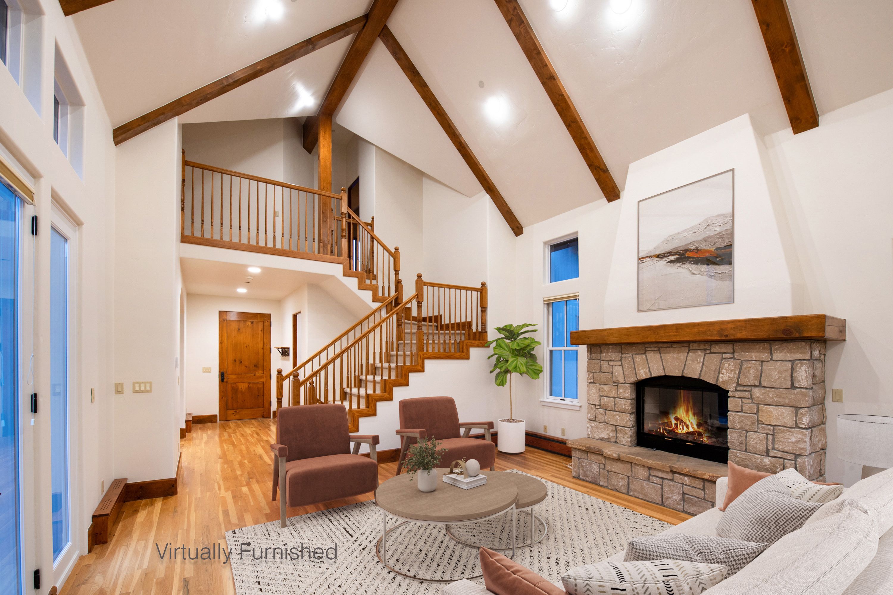 This spacious, high-ceilinged living room features a striking stone fireplace as its focal point, complemented by exposed wooden beams and warm hardwood flooring. The open-concept layout includes a staircase with a wooden railing leading to an upper loft area, creating an airy and inviting atmosphere. Large windows and glass doors allow for ample natural light, highlighting the cozy, virtually furnished seating area.