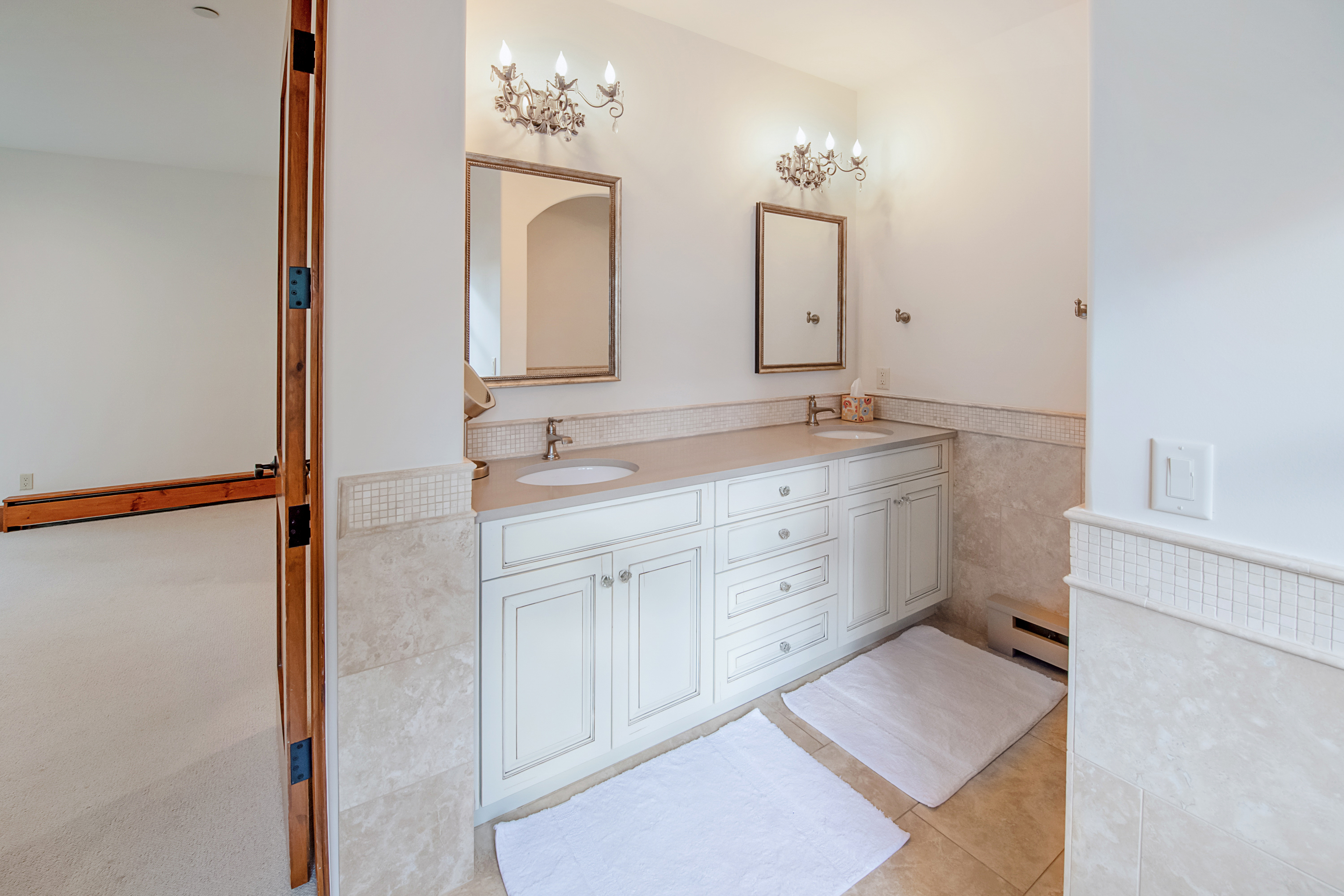 This bright and elegant primary bathroom features a double vanity with white cabinetry, a neutral stone countertop, and dual mirrors illuminated by ornate wall sconces. The space is finished with light-colored tile wainscoting and flooring, creating a clean, cohesive, and sophisticated aesthetic. The perspective captures the vanity area from the doorway, highlighting the functional layout and classic design elements.