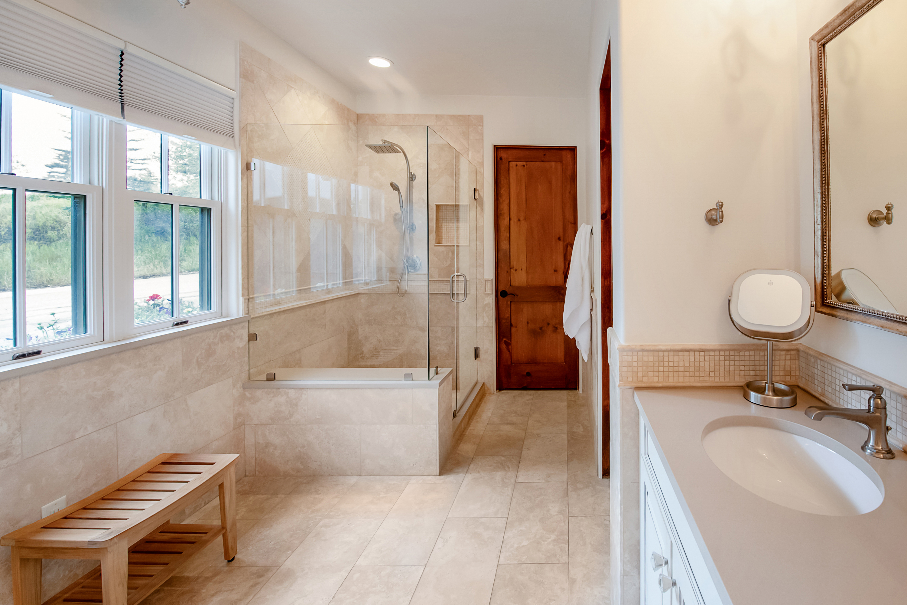 This elegant primary bathroom features a spacious walk-in shower with a glass enclosure, a built-in bench, and neutral stone tile work throughout. A wooden bench sits near the window, while a vanity with a modern sink and a framed mirror completes the sophisticated, spa-like aesthetic. The warm wood door and soft lighting create a welcoming and serene atmosphere.
