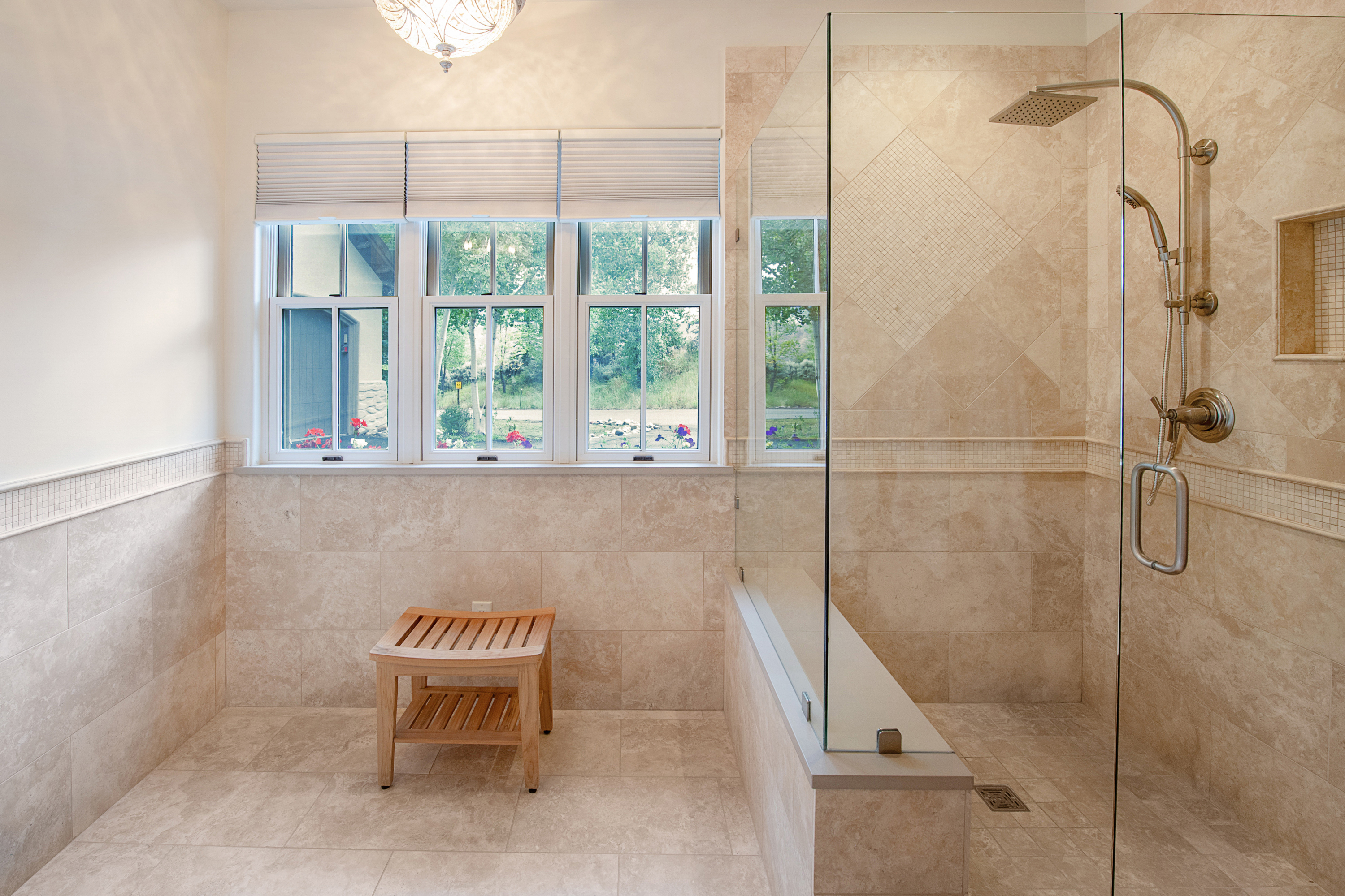 This elegant primary bathroom features a spacious walk-in shower enclosed by a frameless glass panel, accented with neutral-toned travertine tile and a built-in bench. A wooden stool sits in the open area, while large windows with white blinds allow natural light to fill the space, creating a serene and spa-like atmosphere. The design emphasizes high-end finishes and a clean, sophisticated aesthetic.